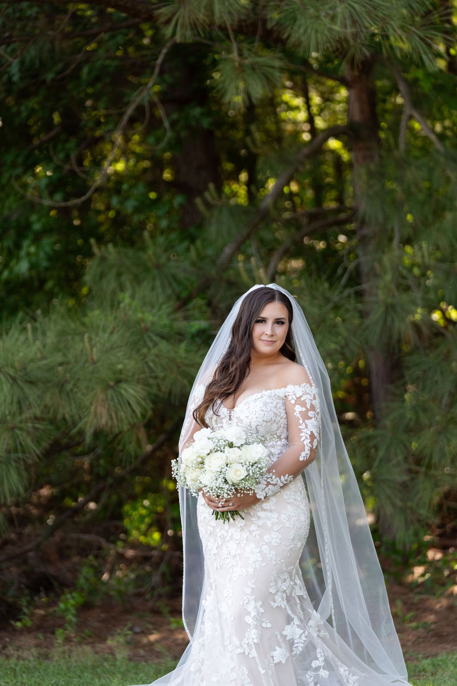 A bride in a white lace wedding dress with long sleeves poses outdoors in a wooded setting holding a bouquet.