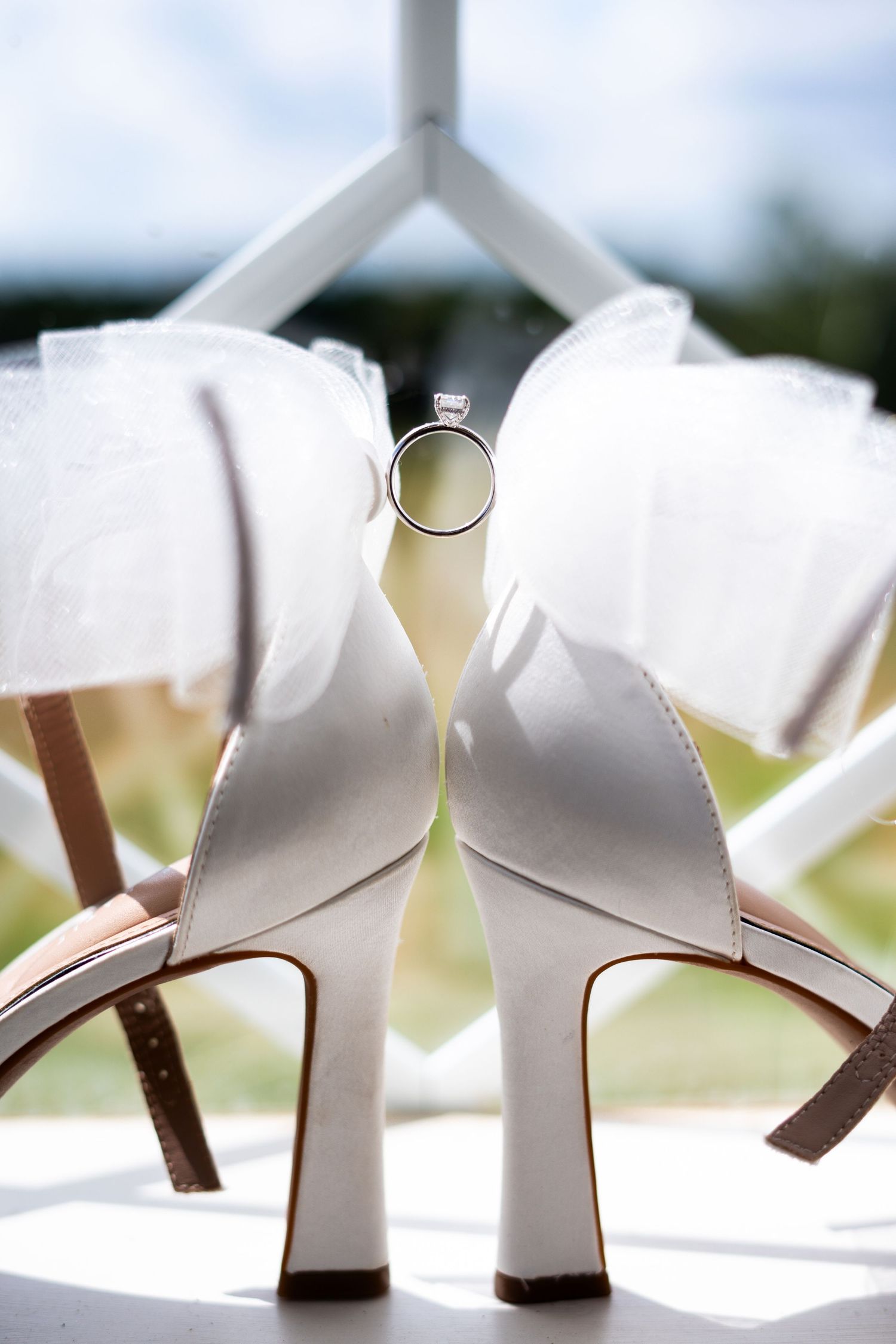 Wedding ring balanced between elegant white bridal heels with a blurred outdoor background.
