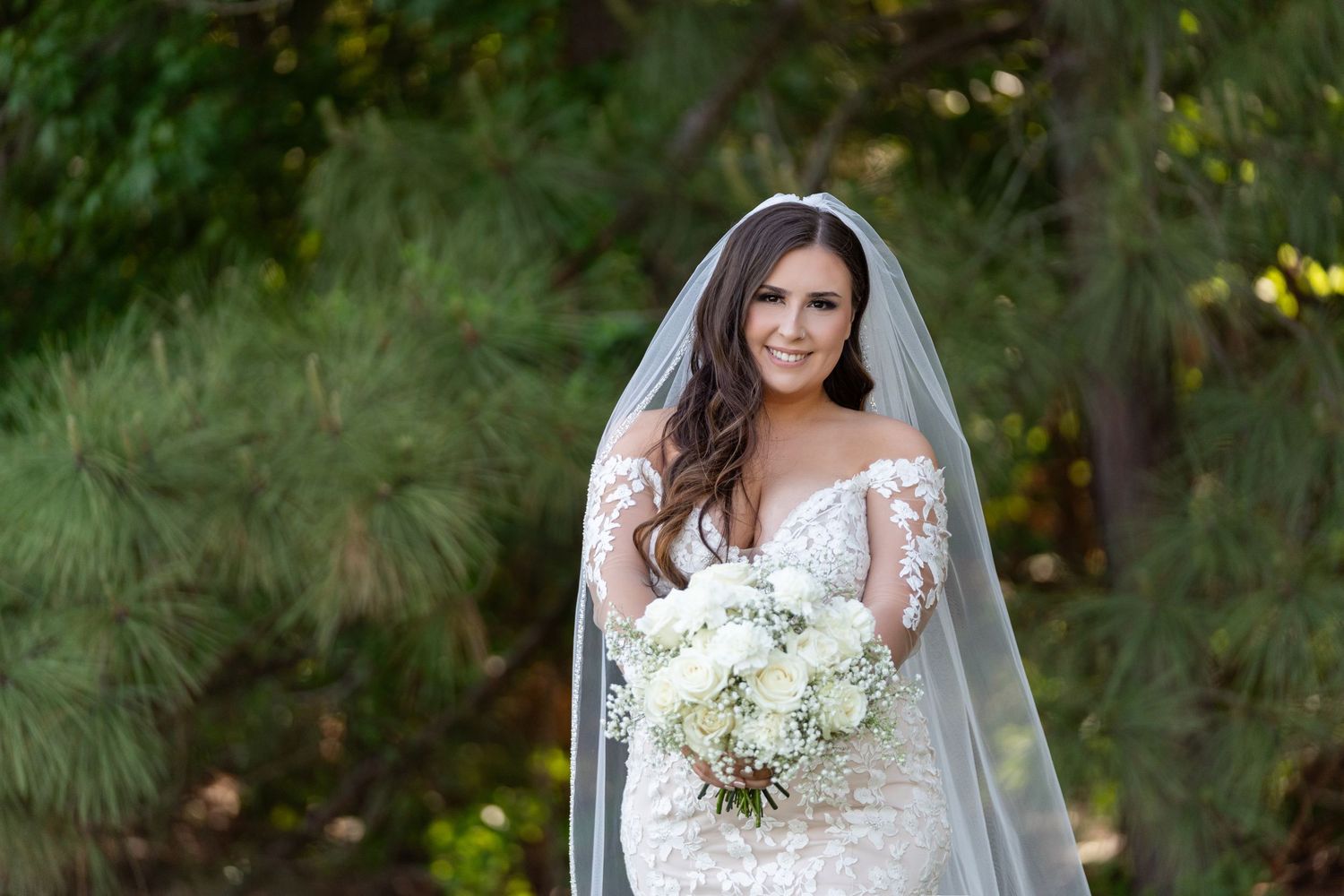 A bride in a lace wedding dress with long sleeves holds a white bouquet in an outdoor garden setting.