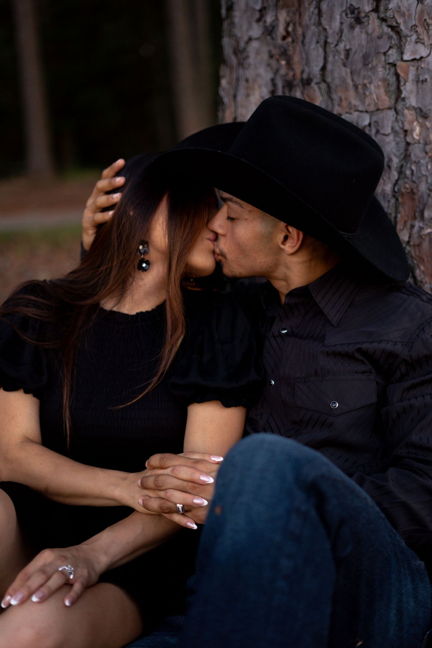 A romantic moment between a couple in black attire sitting by a pine tree.