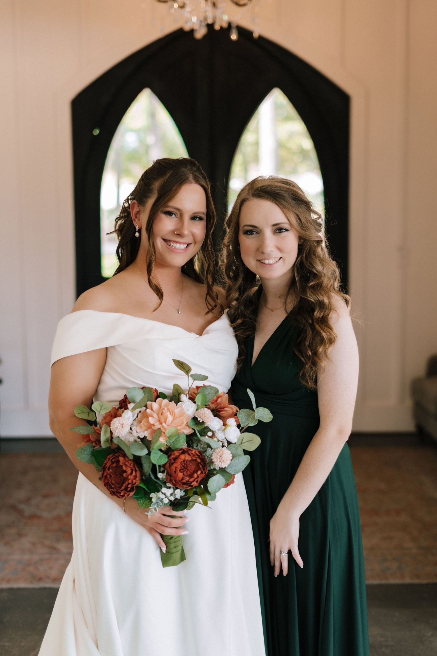 A bride in a white off-shoulder dress holds a peach and burgundy bouquet while posing with a bridesmaid in emerald green.