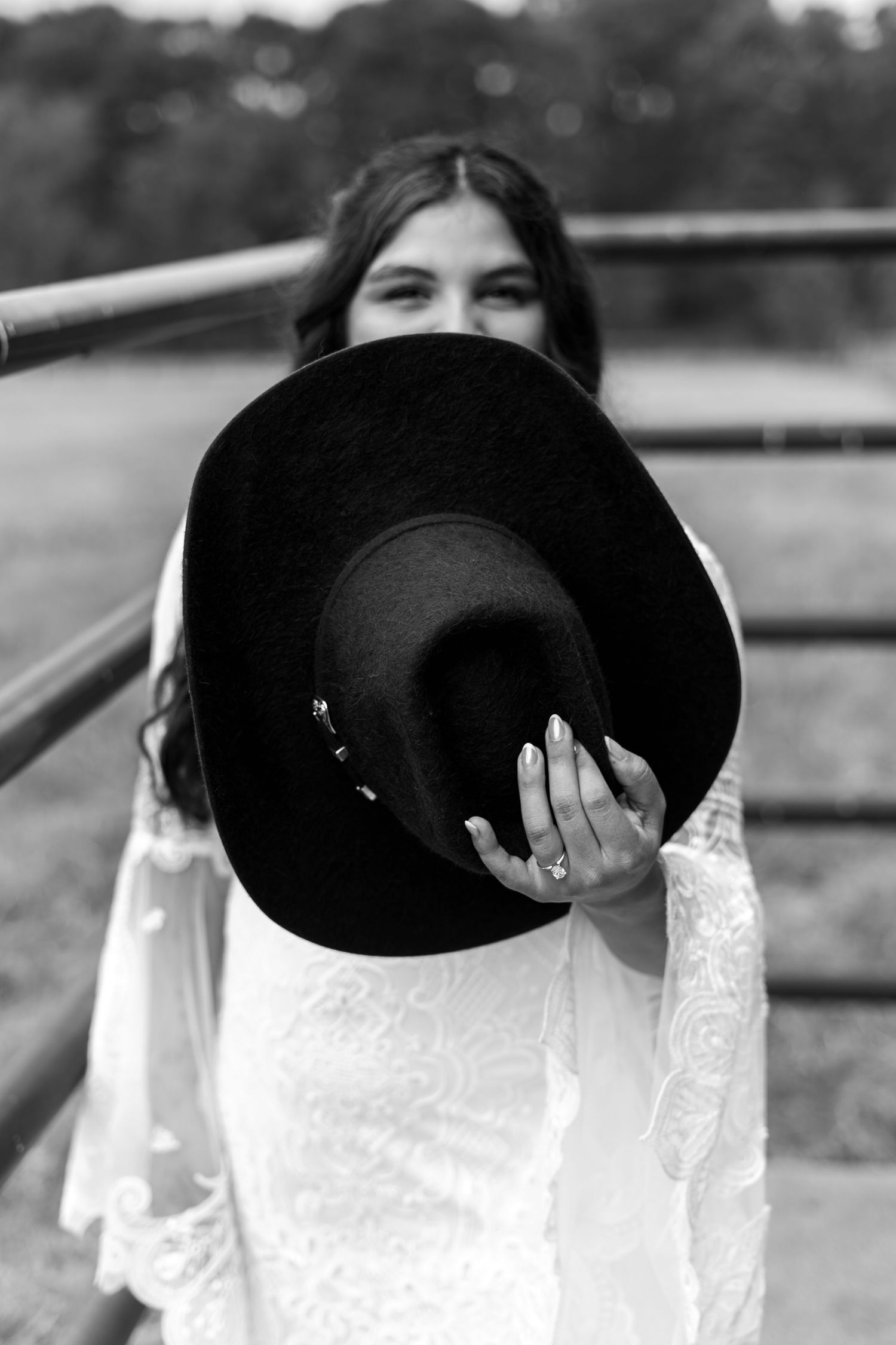A black and white portrait of someone holding a black hat in front of their face while wearing a white lace dress.