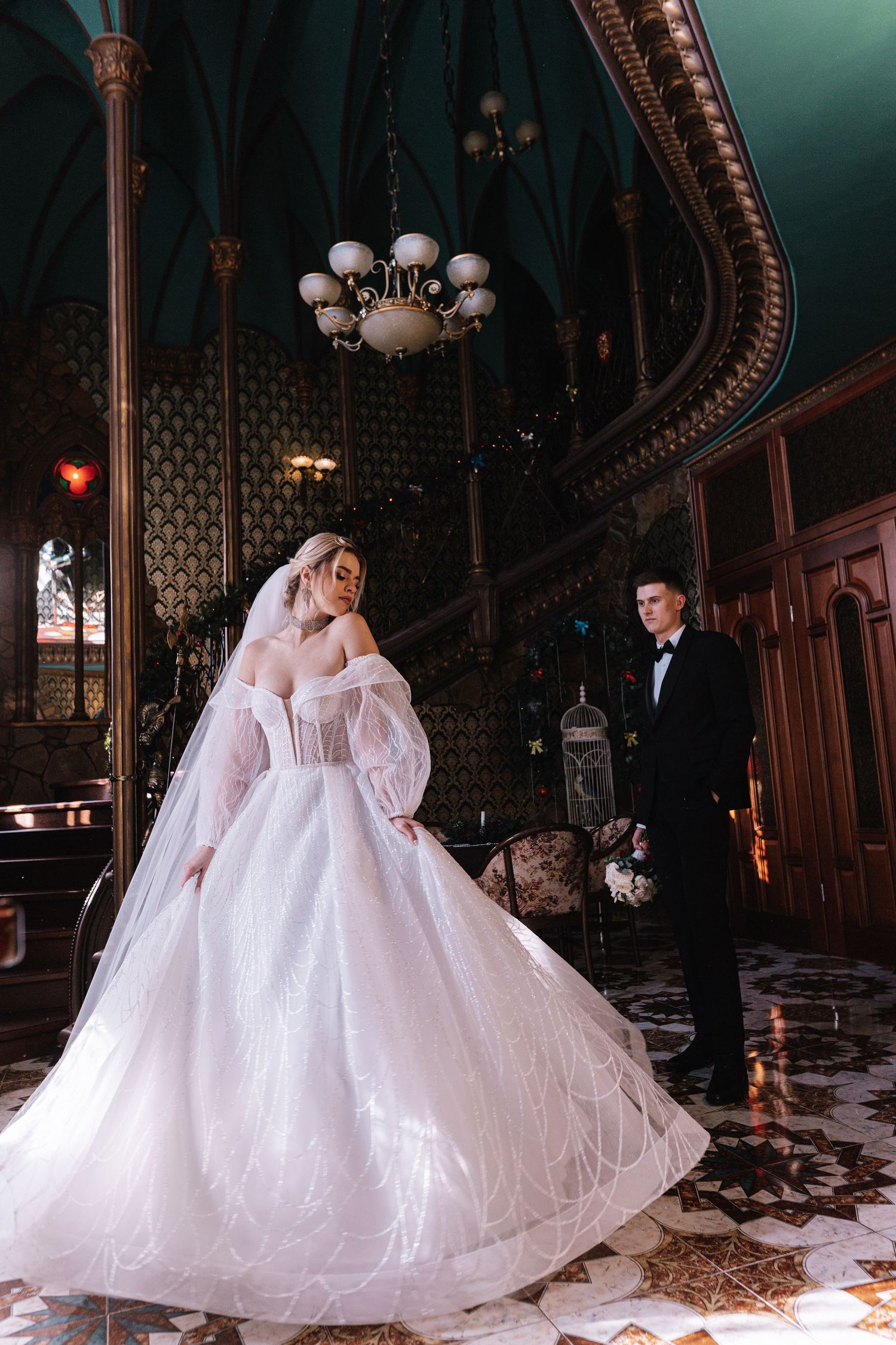 A couple in formal wedding attire dance together in an ornate vintage ballroom with elegant chandeliers and architectural details.