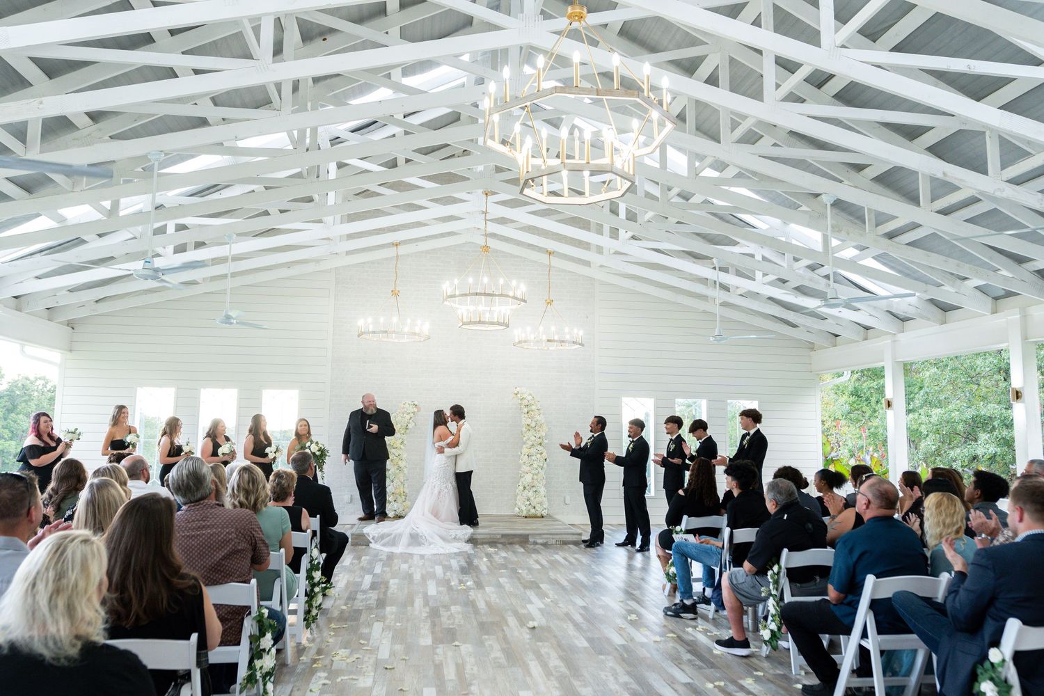 Wedding ceremony taking place in a modern white venue with exposed ceiling beams and rows of seated guests.
