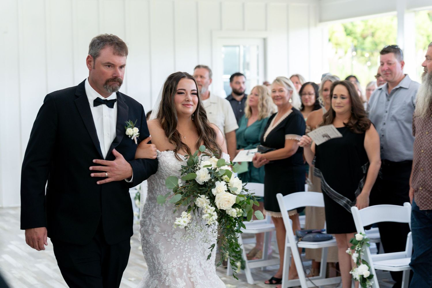A couple walks down the aisle during their outdoor wedding ceremony with guests looking on from white chairs.