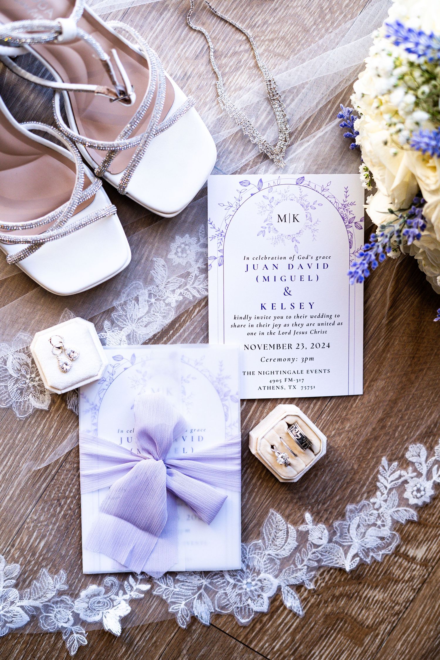 Wedding invitation suite in lavender and white with lace details and floral accents on rustic wooden surface.