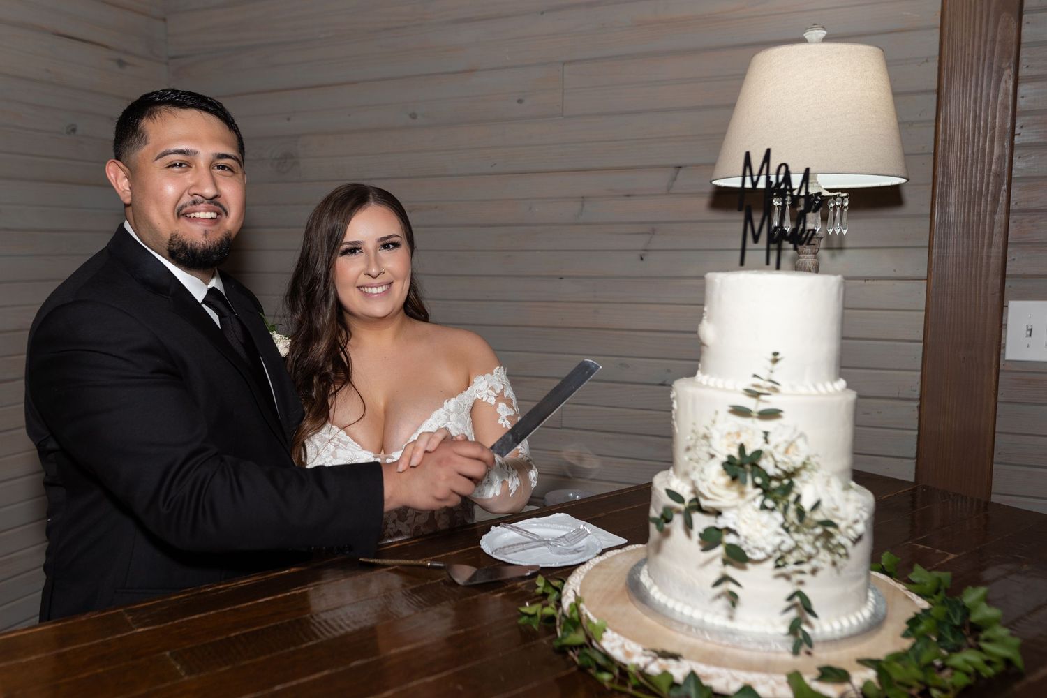 A couple cuts into their elegant four-tier white wedding cake adorned with greenery at a rustic wooden table.