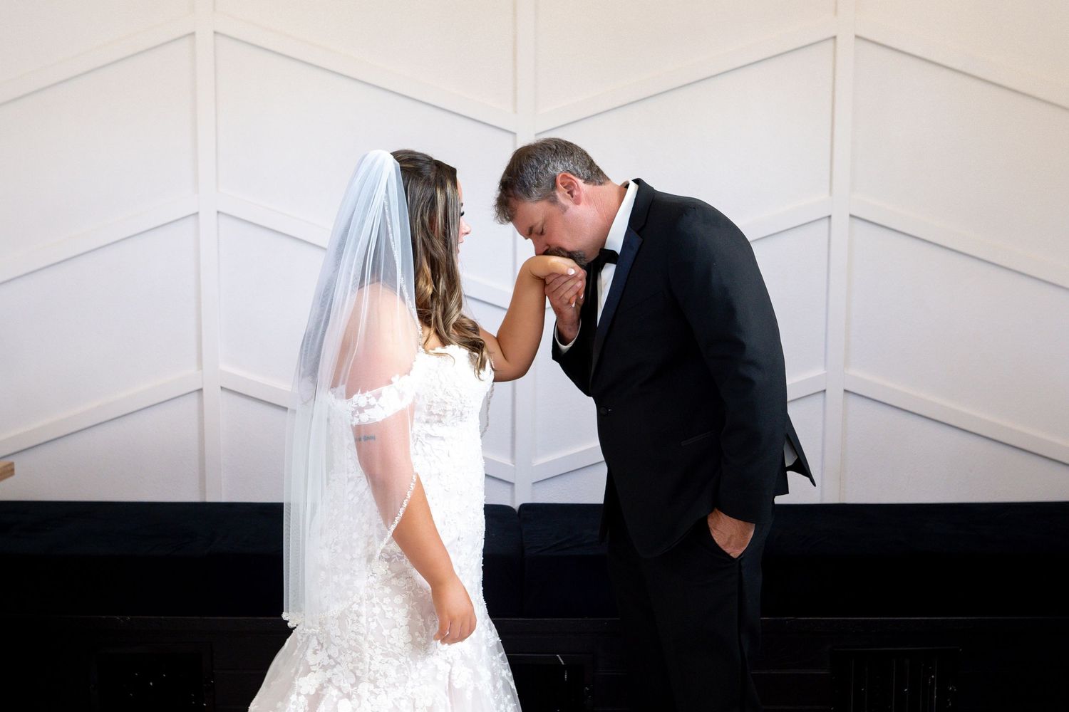 A bride in a white lace gown and veil shares a tender moment with someone in a black suit against a white wall.