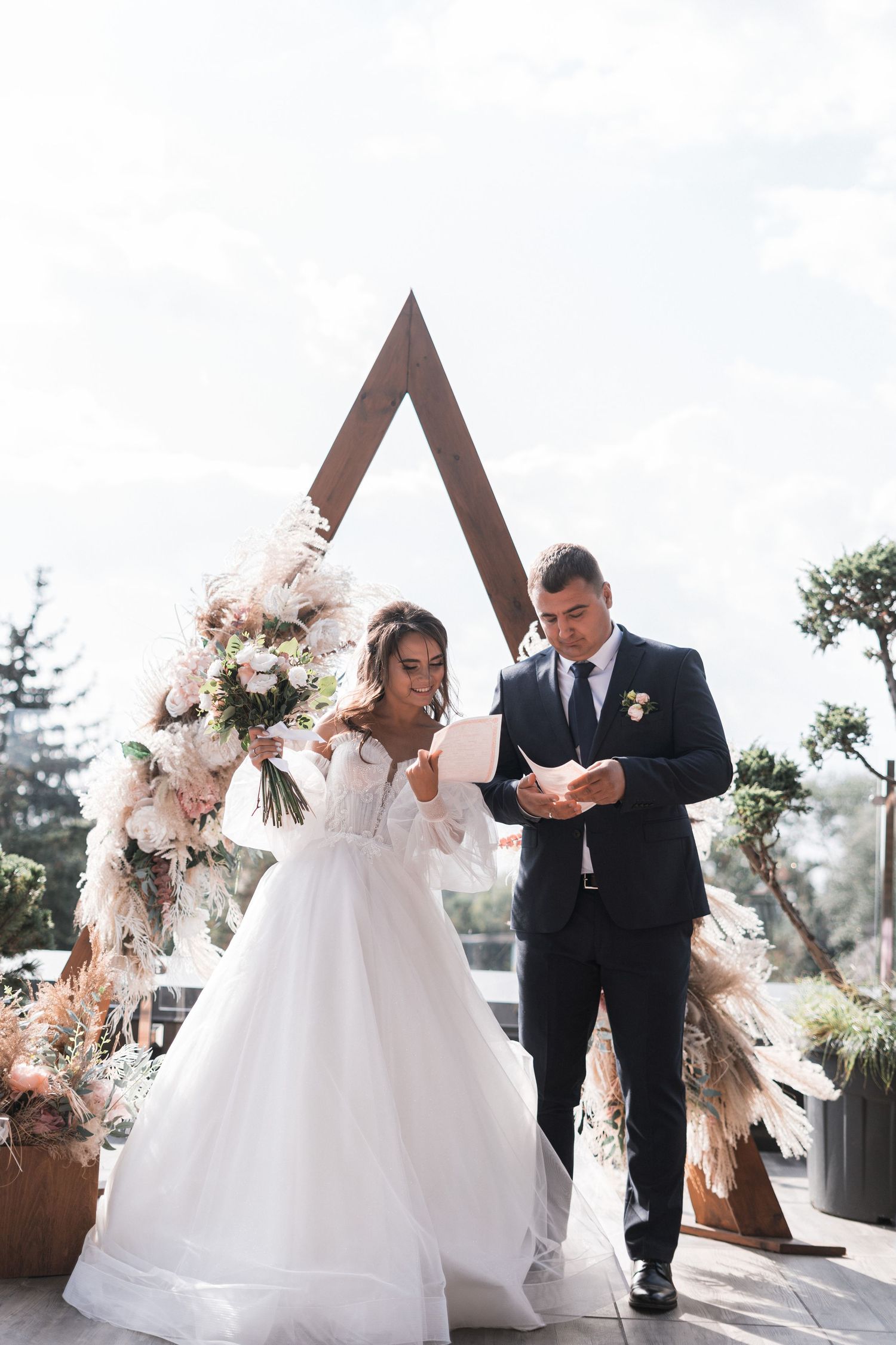 An emotional outdoor wedding ceremony with a wooden arch and floral arrangements in soft natural light.