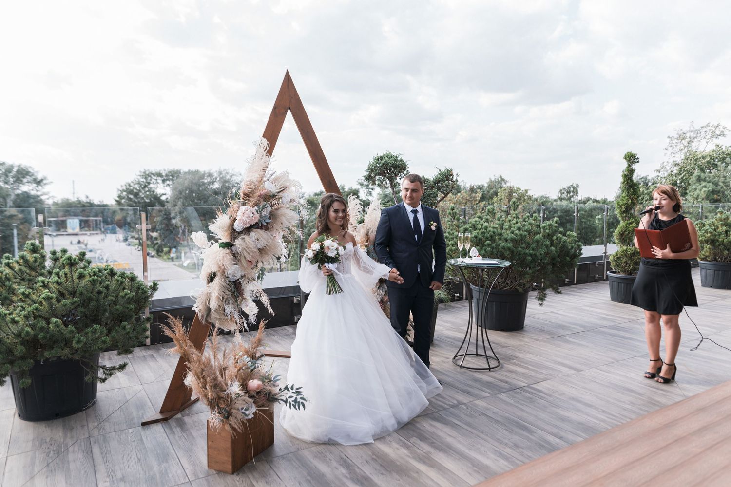 A wedding ceremony takes place on a modern rooftop deck decorated with potted plants and a geometric wooden arch.