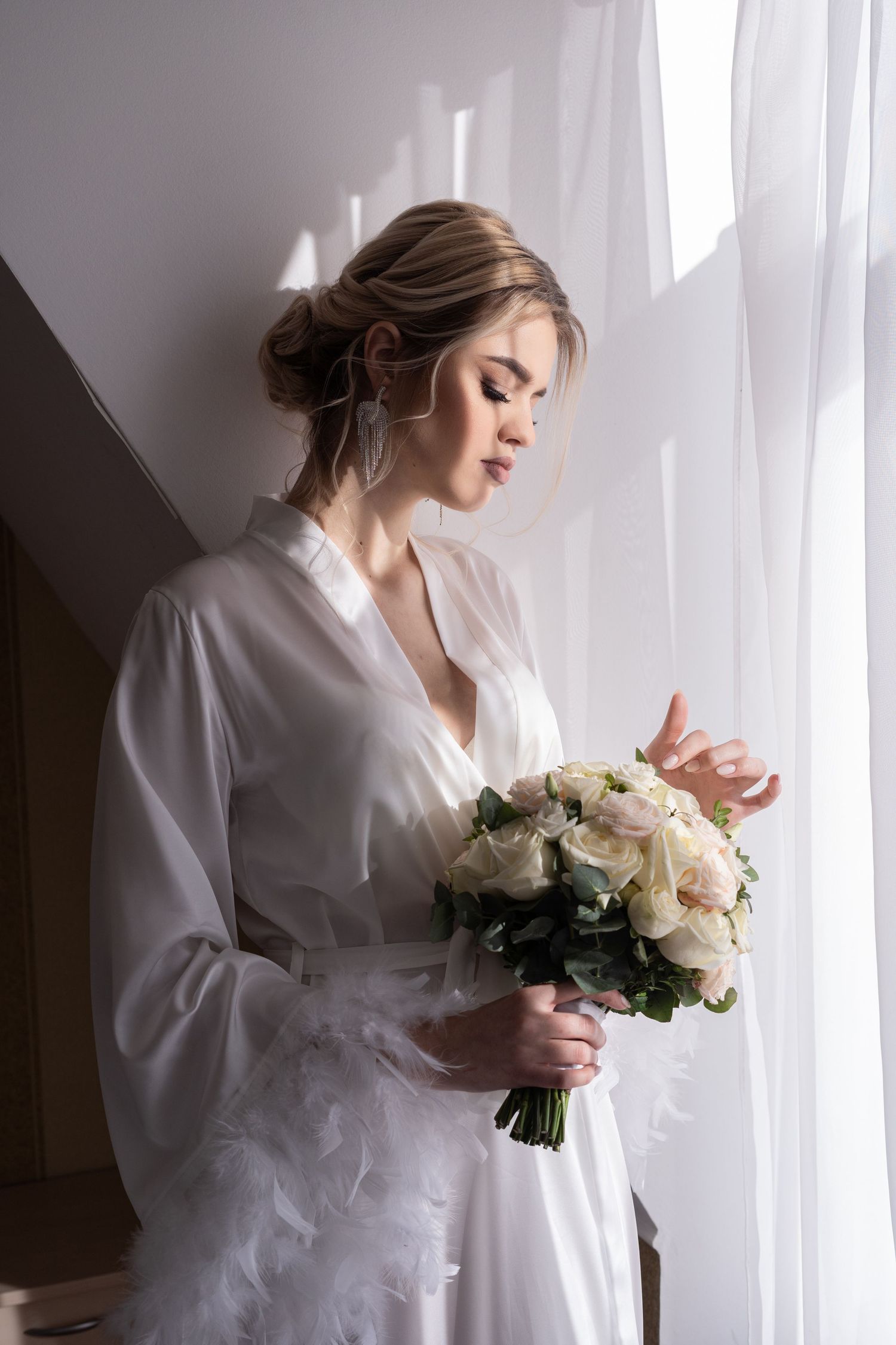 A figure in a white robe holds a delicate bouquet of white roses near a sunlit window.