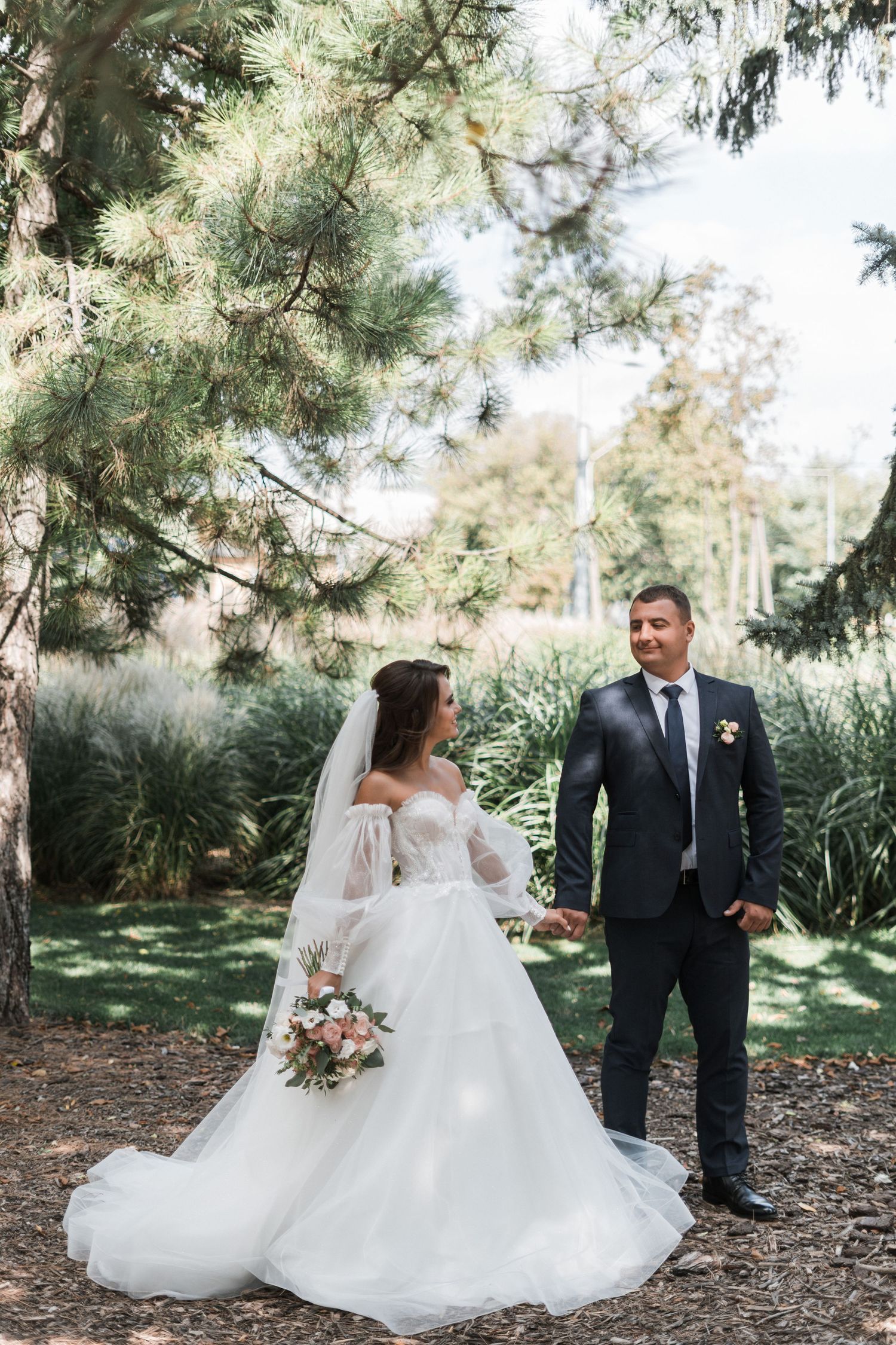 A bride in a flowing white gown and groom in a dark suit pose together in a lush garden setting.