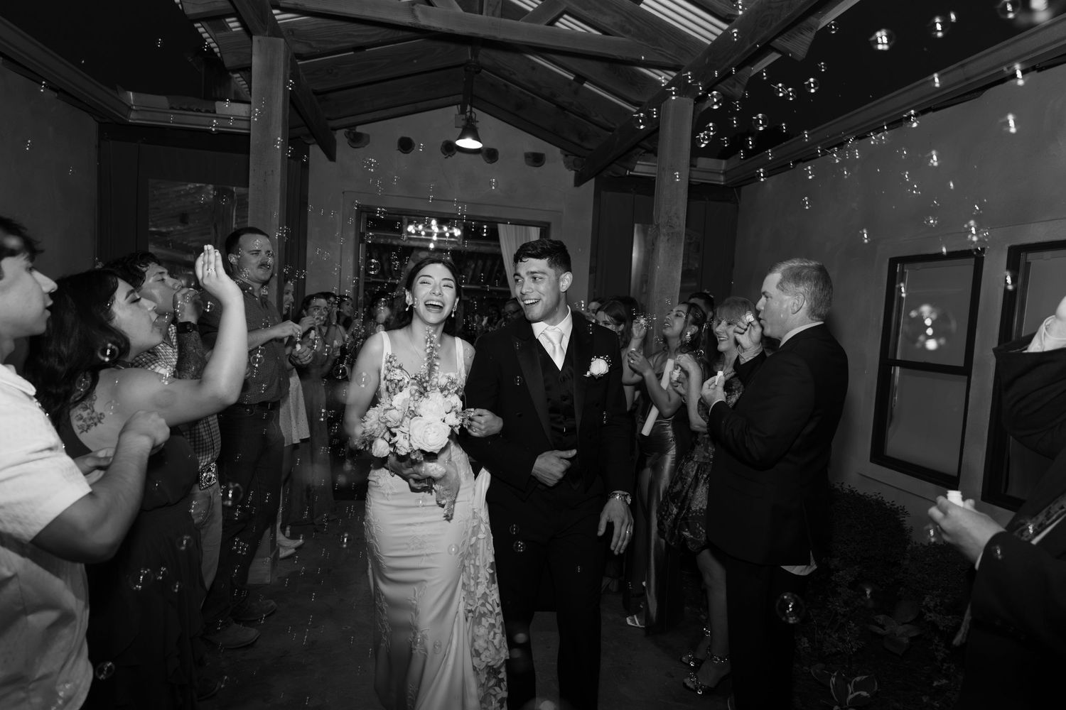 Bride holding a bouquet and groom walk arm in arm through a cheering crowd of wedding guests as bubbles float in a rustic indoor venue.