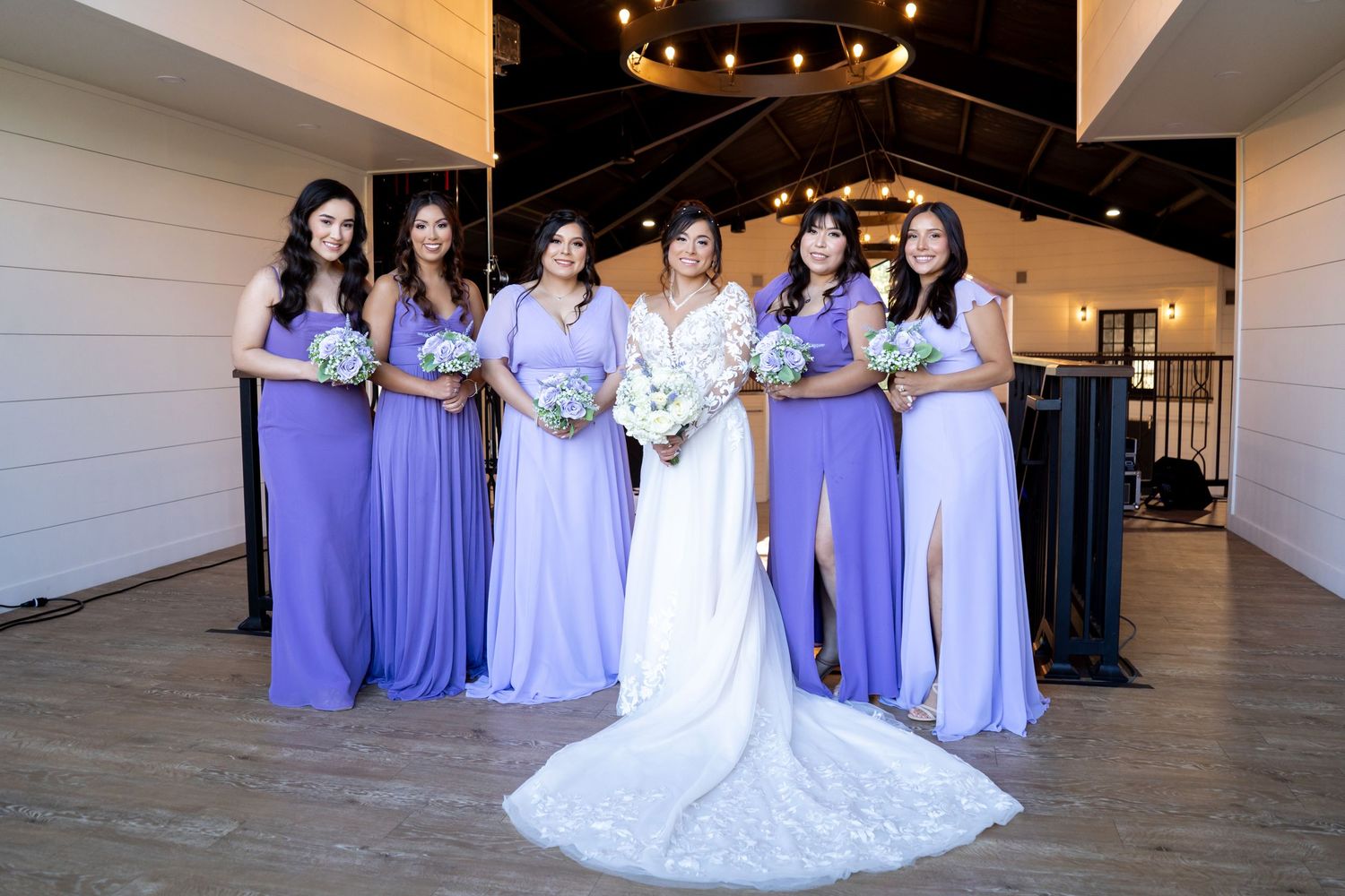Wedding party in purple bridesmaid dresses and white gown posed in elegant venue with string lights.