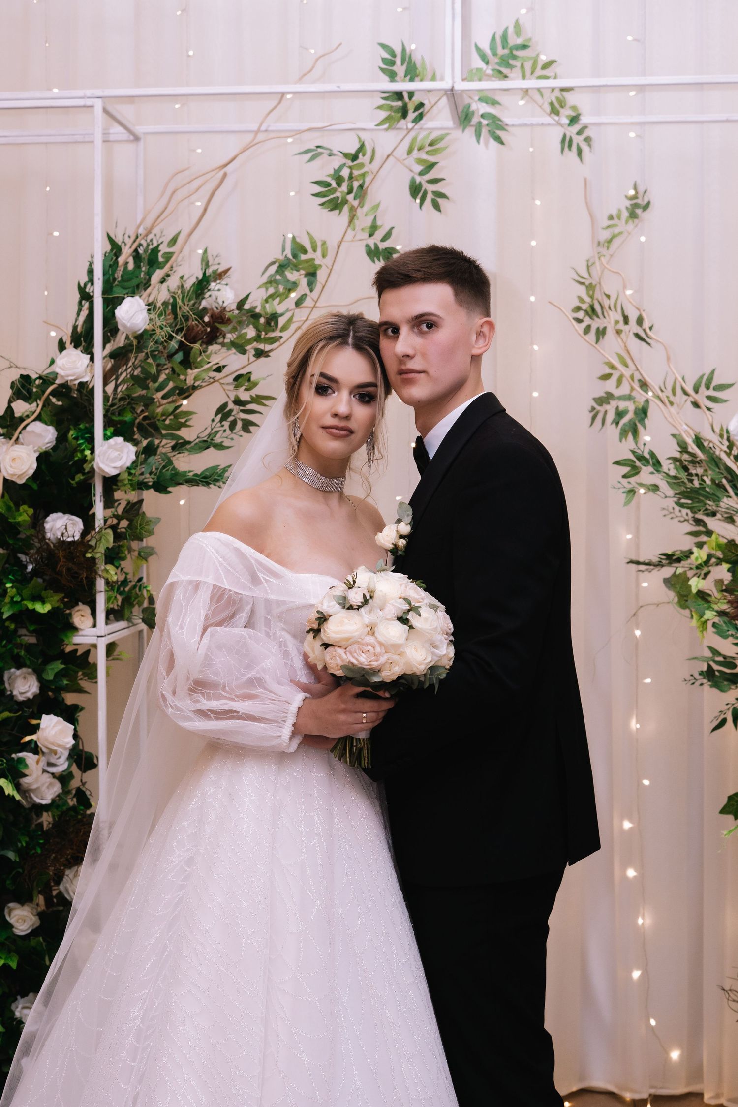 A romantic wedding scene with string lights and greenery decor as newlyweds pose together in traditional attire.