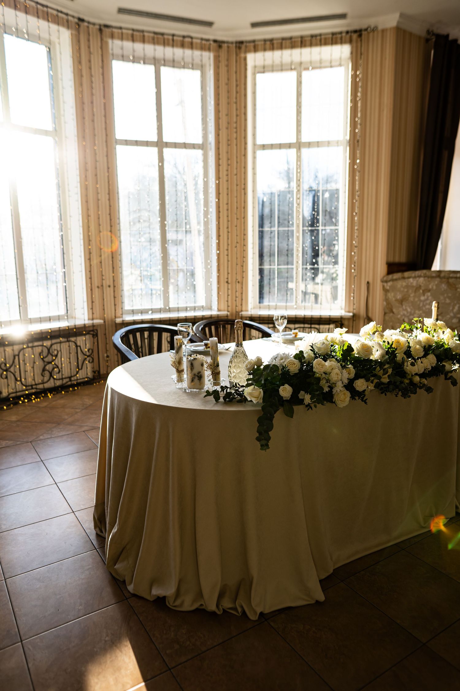 Round banquet tables with elegant white tablecloths arranged in a bright room with large windows and floral centerpieces.