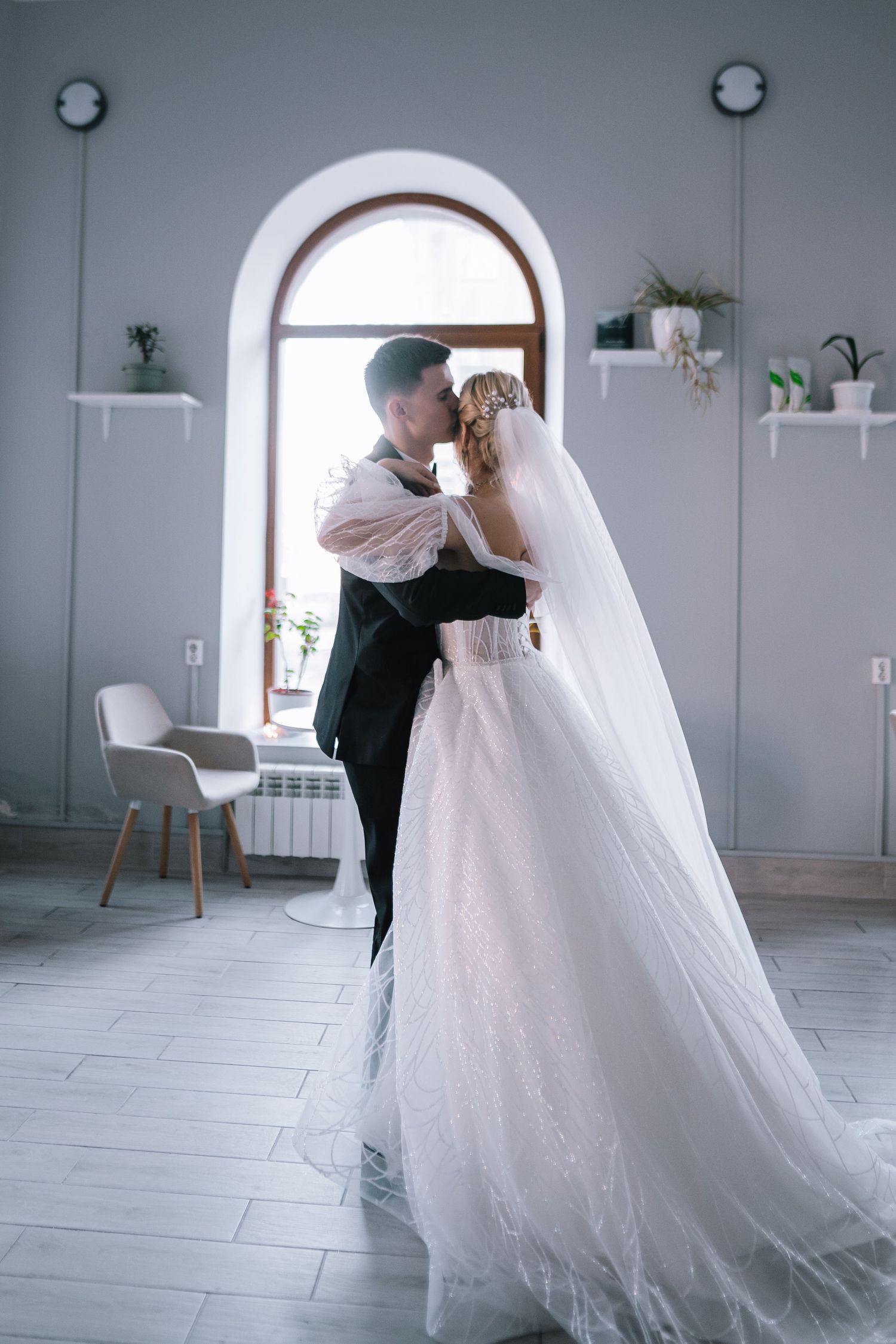 A romantic wedding couple shares an intimate dance moment in a bright, minimalist studio space with arched windows.