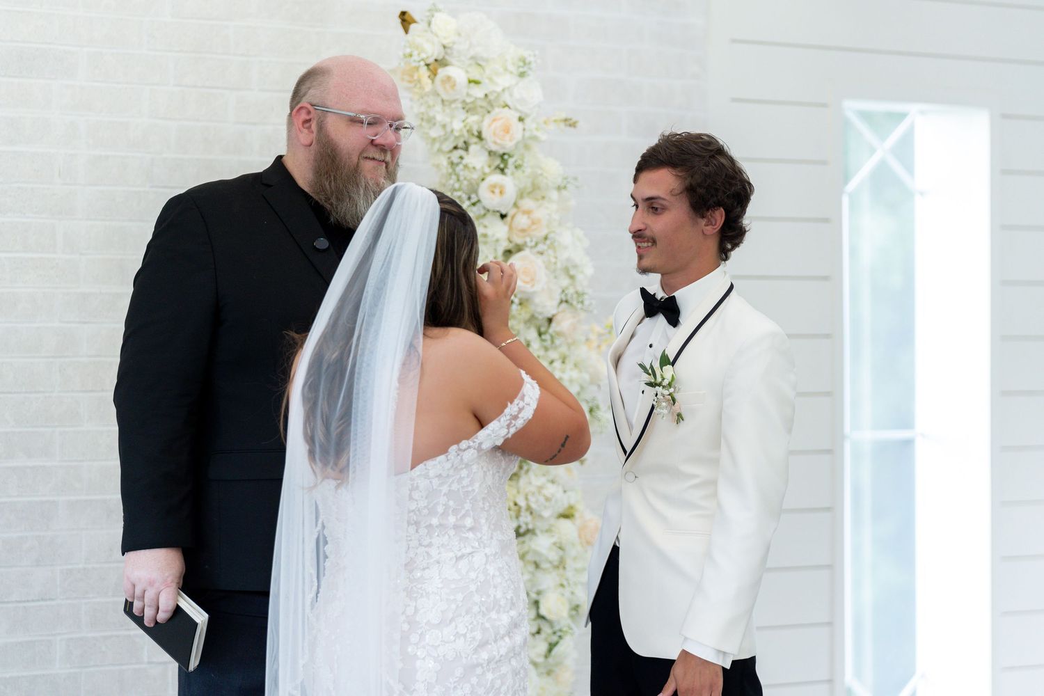 A wedding ceremony takes place in front of a white floral arch with the bride wearing a veil and groom in white tuxedo.