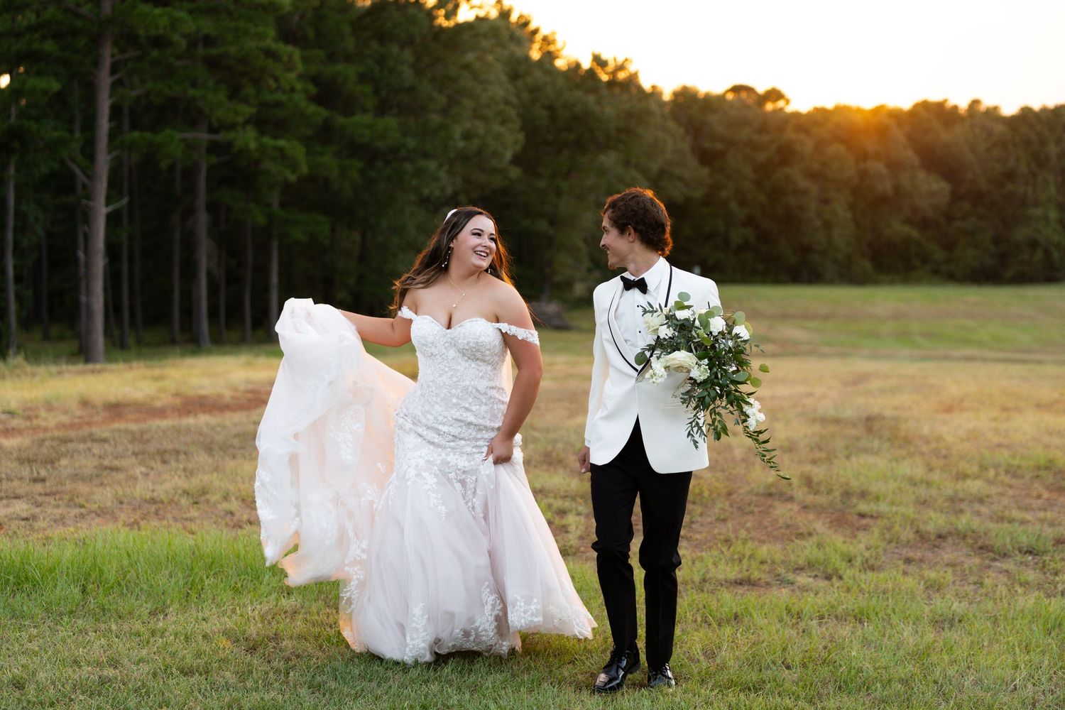 A couple in wedding attire walks hand in hand through a golden sunlit field at sunset.