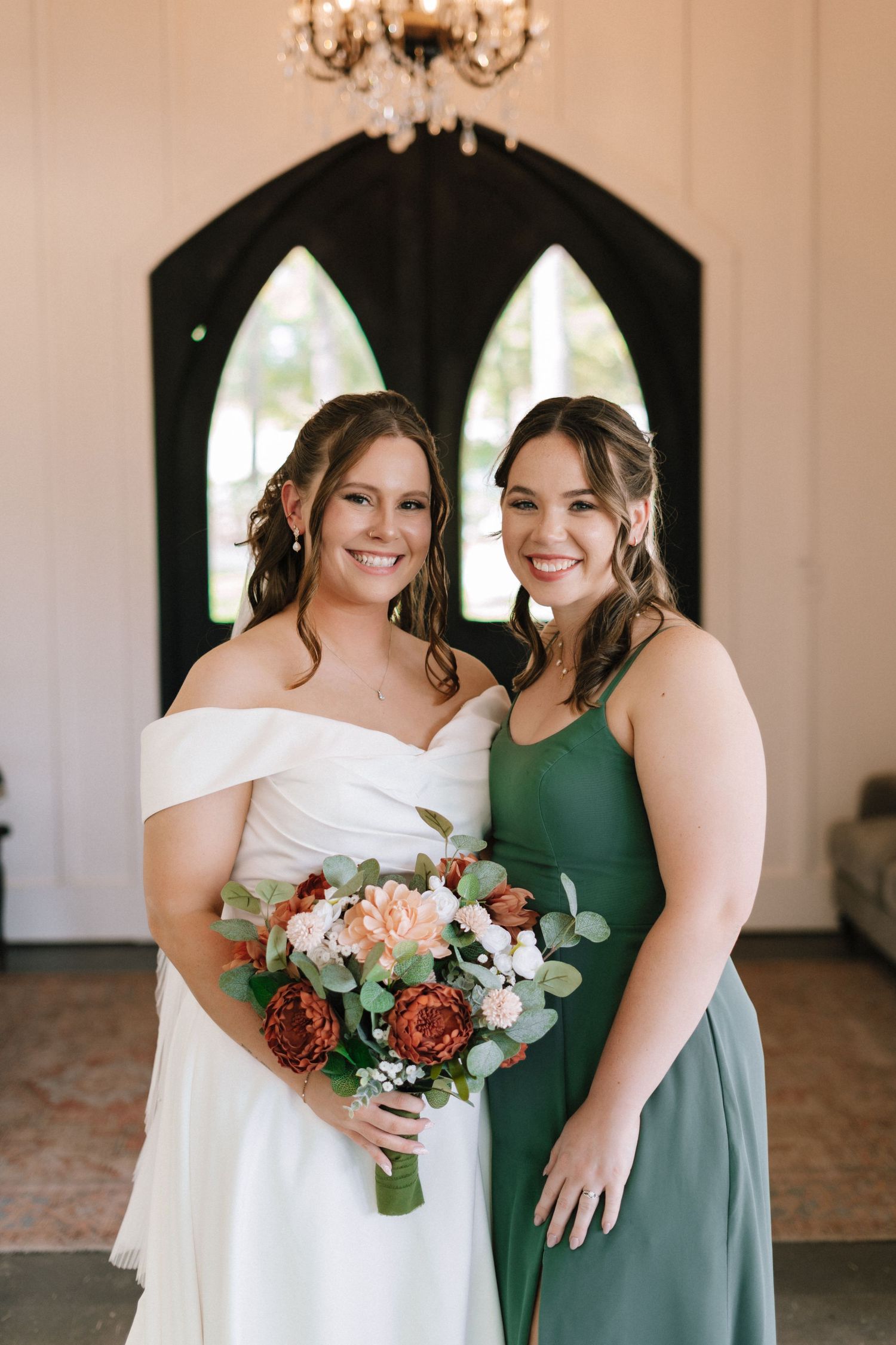 Two women in elegant wedding attire share joyful moments while holding a beautiful bouquet of pink and red flowers.