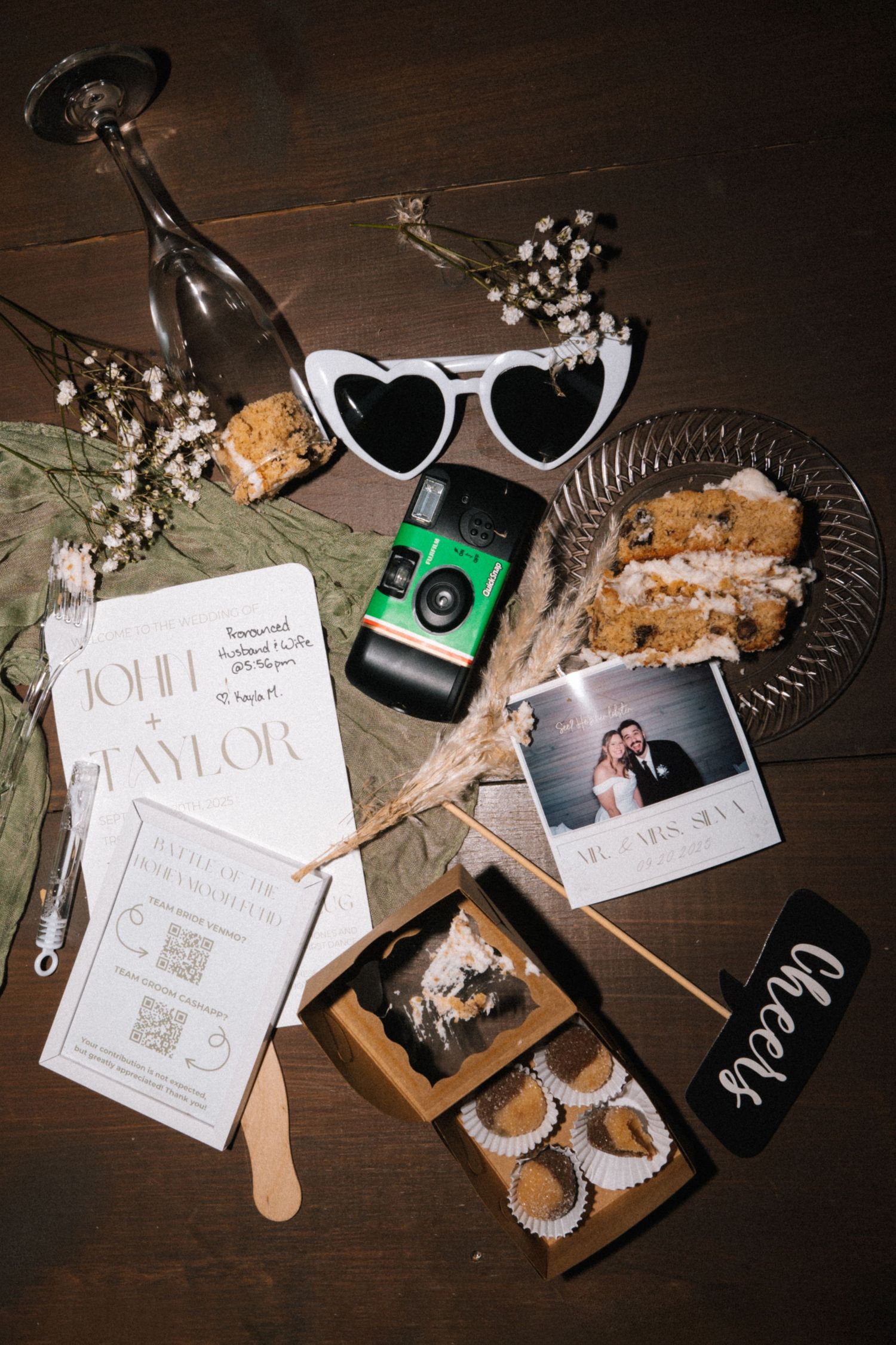 Wedding detail flatlay with invitations, sunglasses, and decorative elements on dark wooden surface.