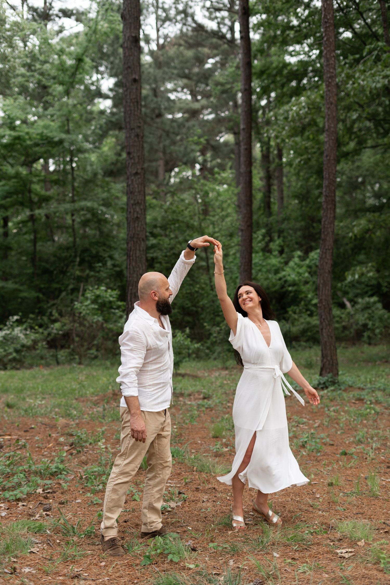 A couple in white clothing dances together in a forest clearing.