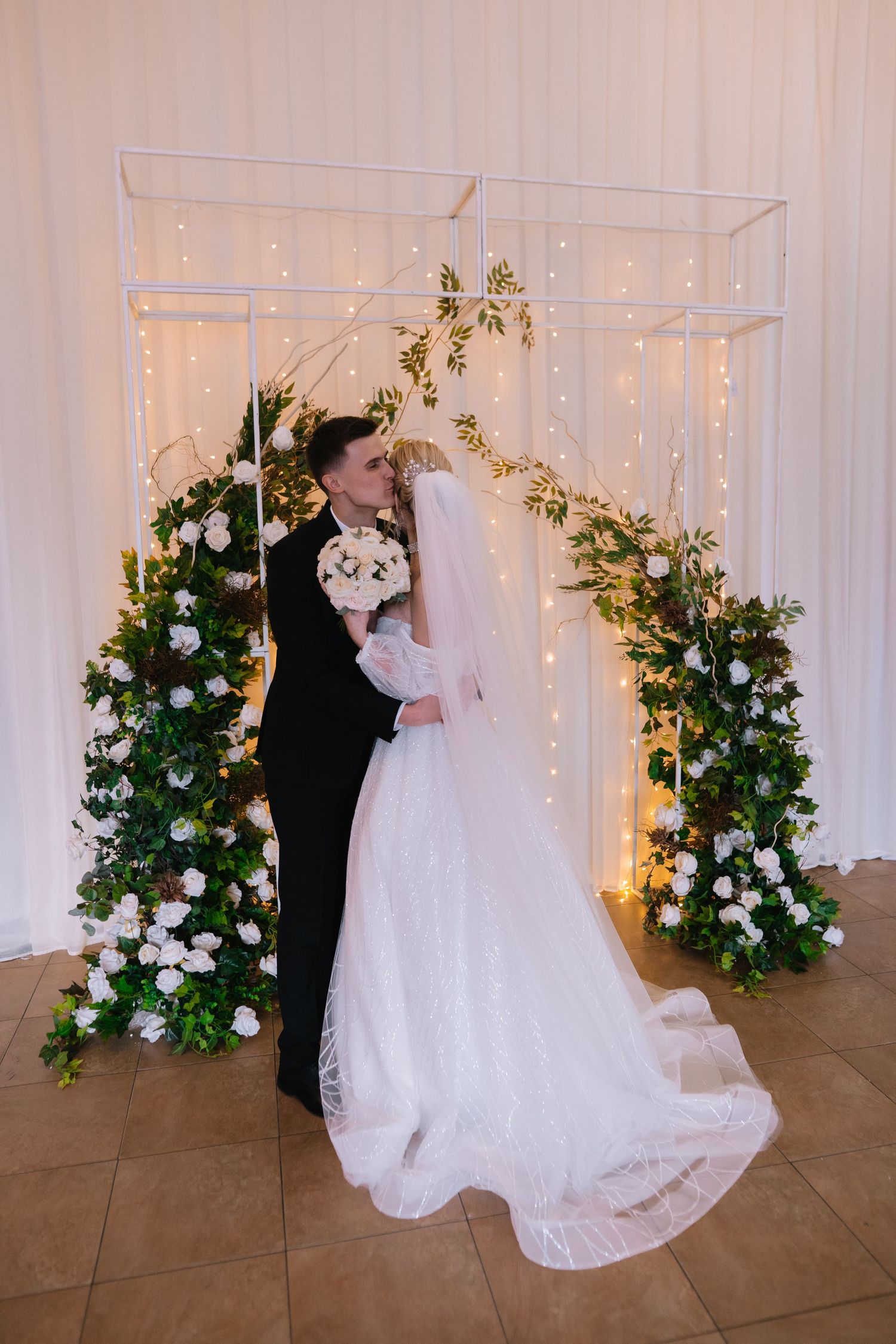 A romantic wedding couple shares their first dance surrounded by elegant floral arrangements and white drapery.