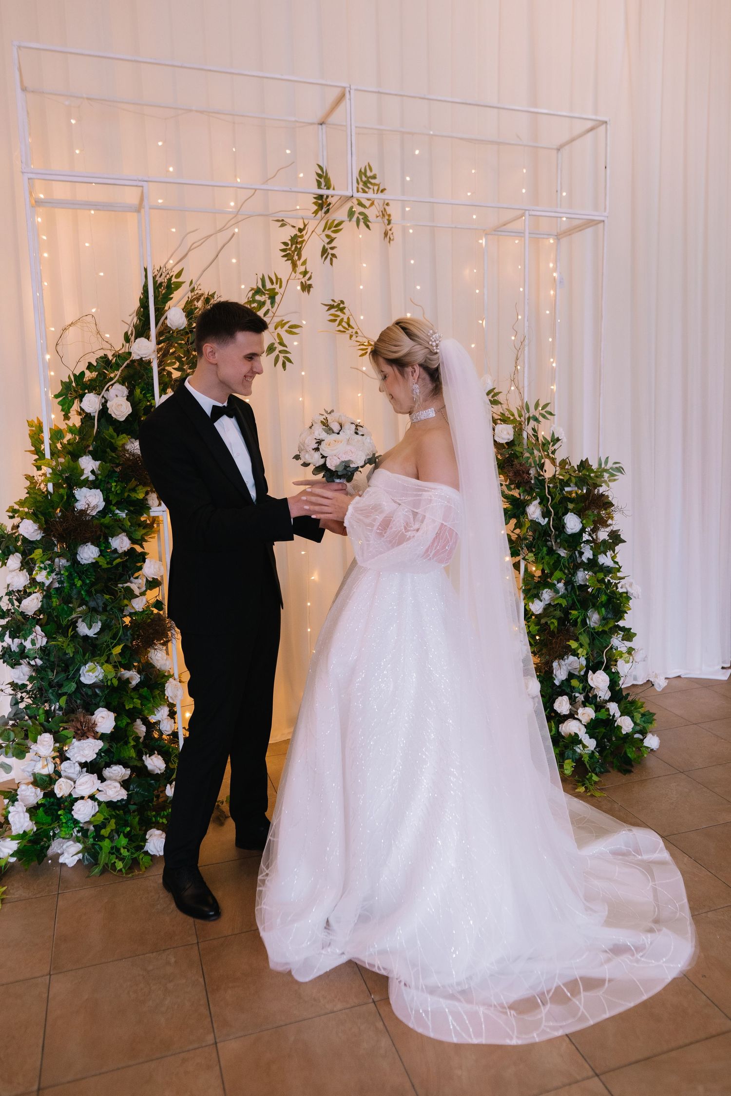 A couple dances together at their wedding reception, with white floral arrangements and twinkling lights in the background.
