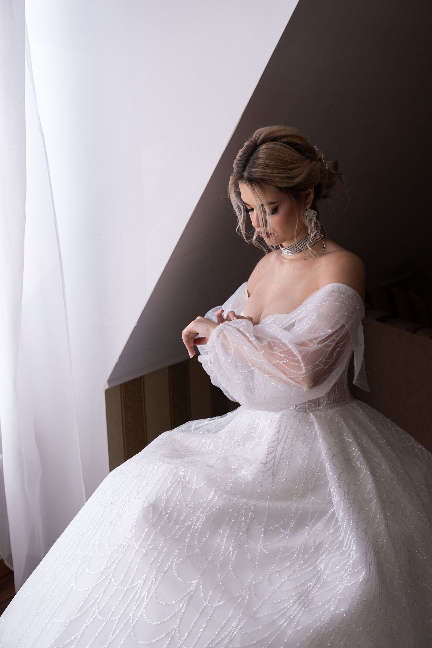 A bride in a flowing white gown sits gracefully by a window with sheer curtains in a romantic boudoir setting.