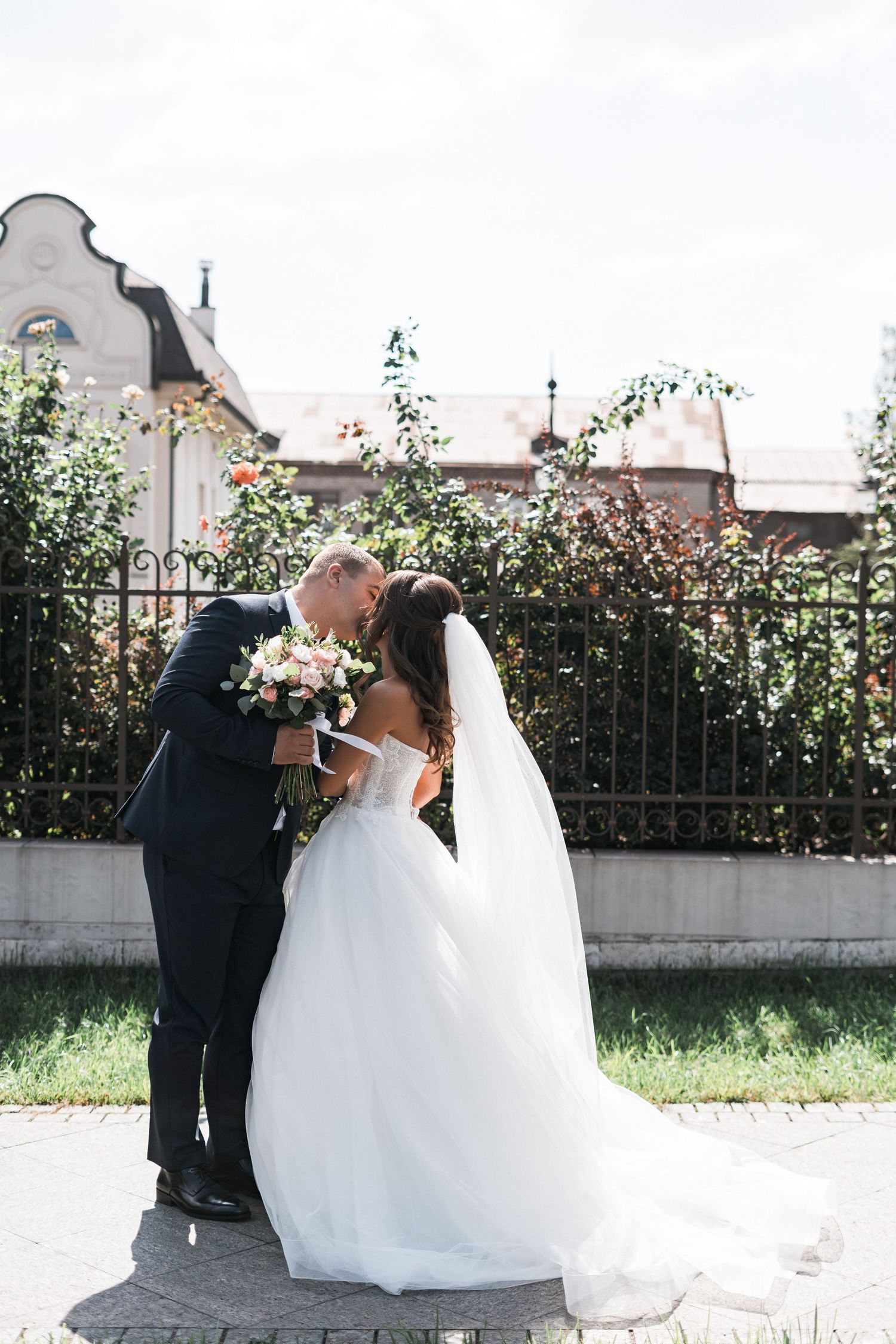 A bride and groom share a romantic moment in front of an iron fence and flowering garden on their wedding day.