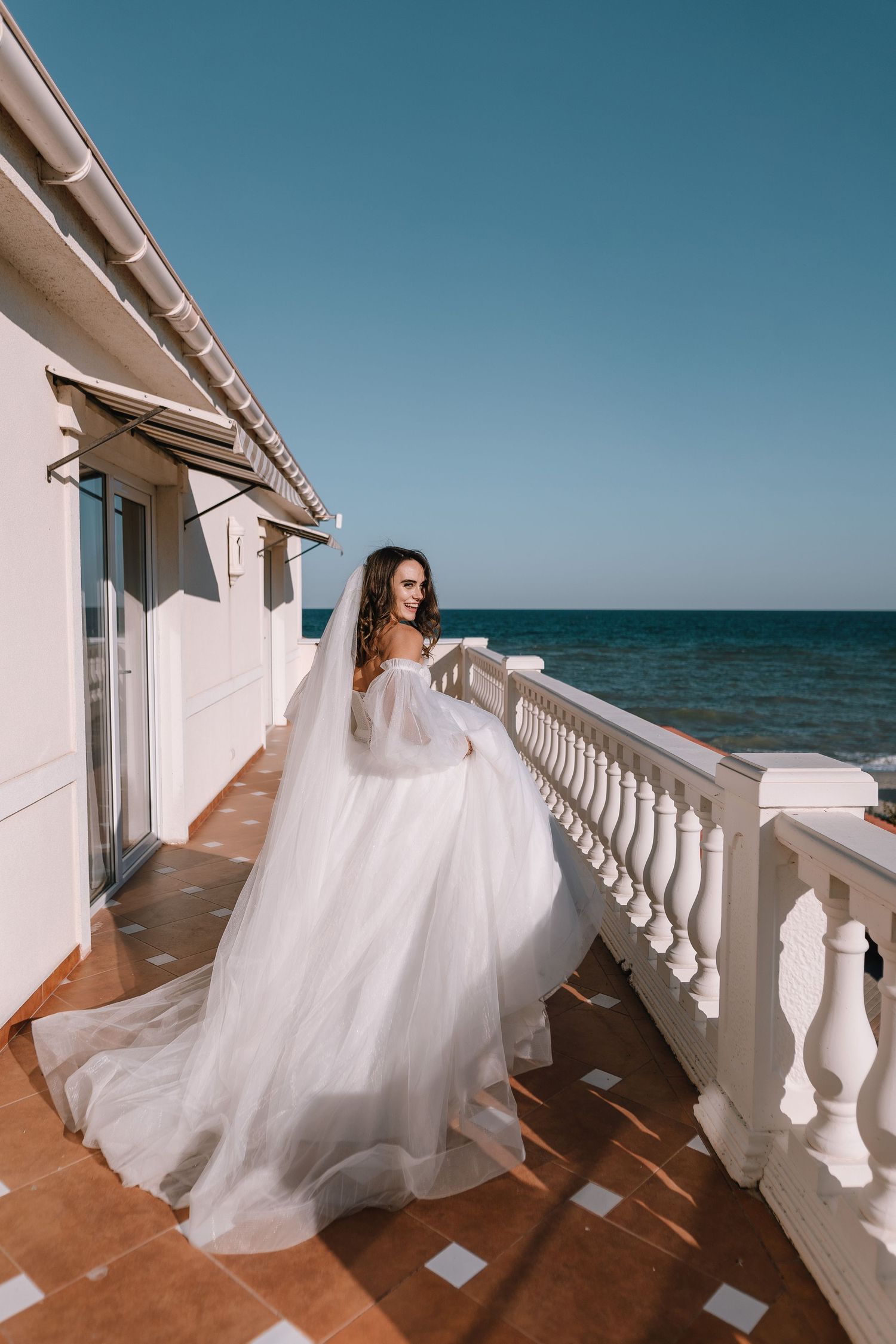 A flowing white wedding dress billows in the ocean breeze on a seaside terrace with white balustrades.
