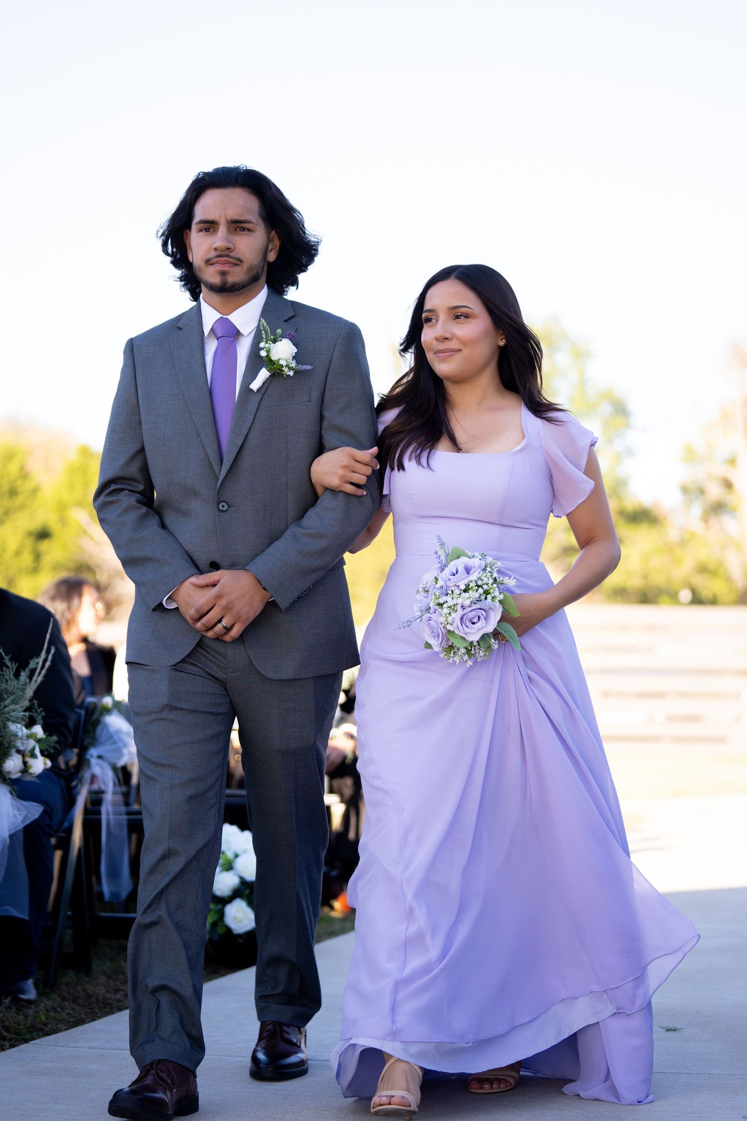 Wedding party members in purple dresses and gray suits walking during outdoor ceremony.
