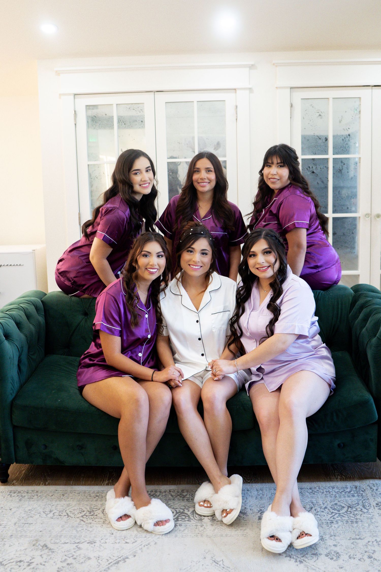 Wedding party members in purple dresses and white robes pose together on emerald velvet sofa.