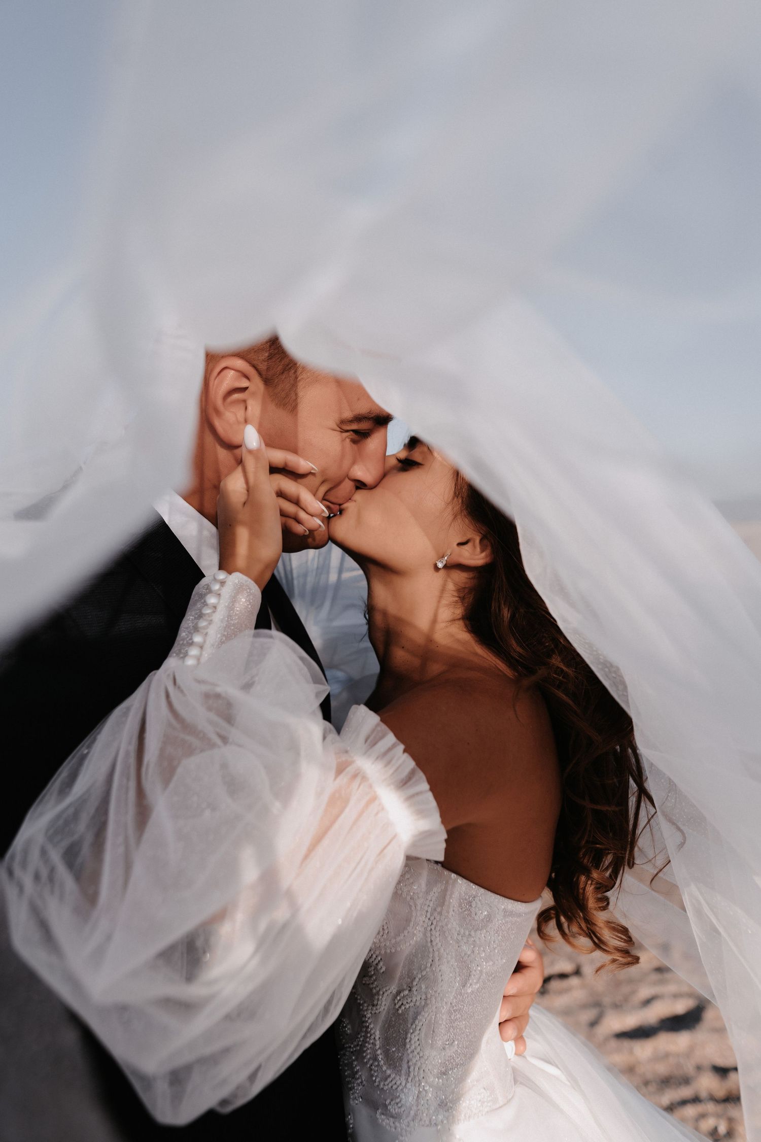 A romantic moment between newlyweds under a flowing white veil against a cloudy sky.