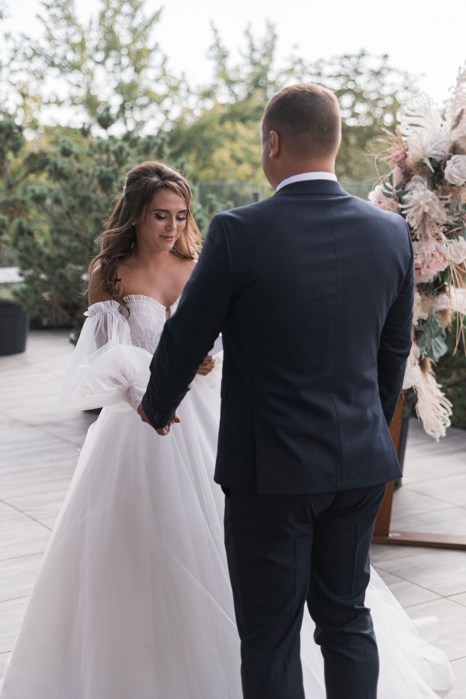 A couple shares a romantic moment during their wedding ceremony next to a floral arch on an outdoor patio.