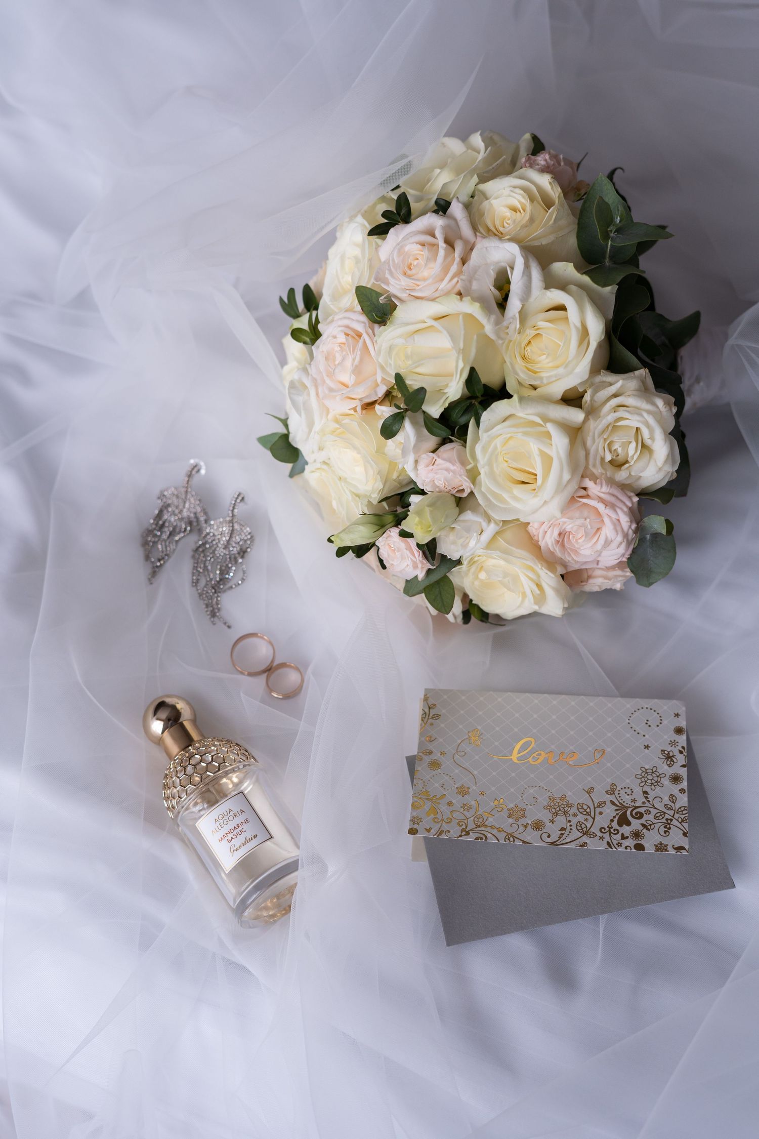 Elegant wedding details featuring cream roses, gold rings, and delicate tulle fabric arranged on white background.