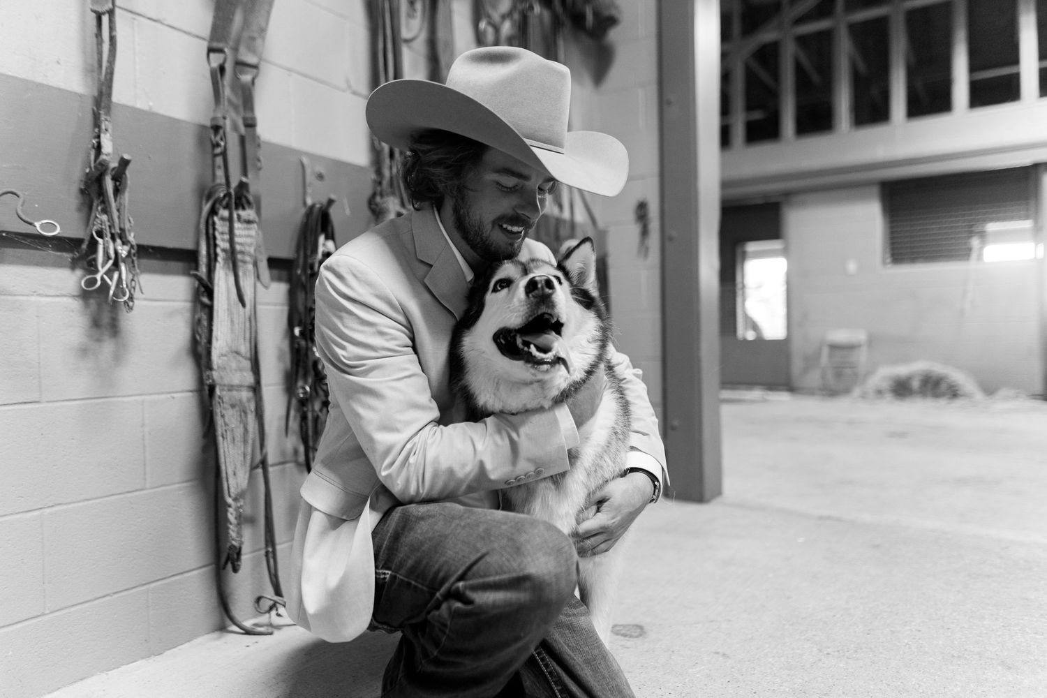 A black and white photo showing a person in a cowboy hat hugging a dog in a rustic setting.