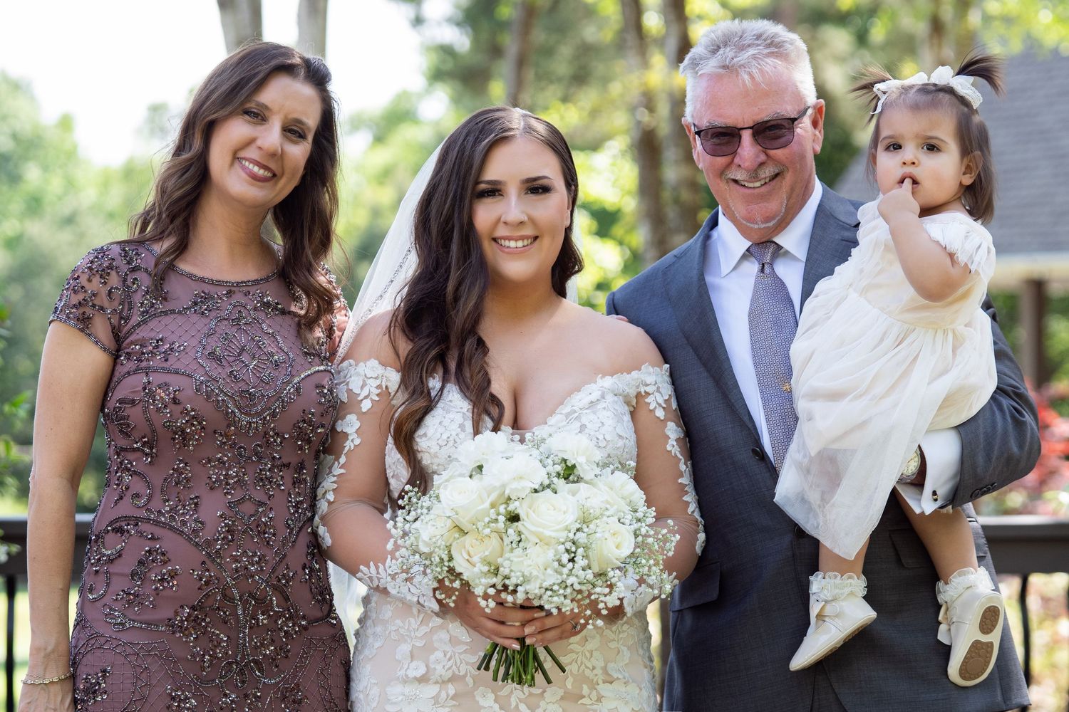 A group poses together at an outdoor wedding celebration with a white floral bouquet.