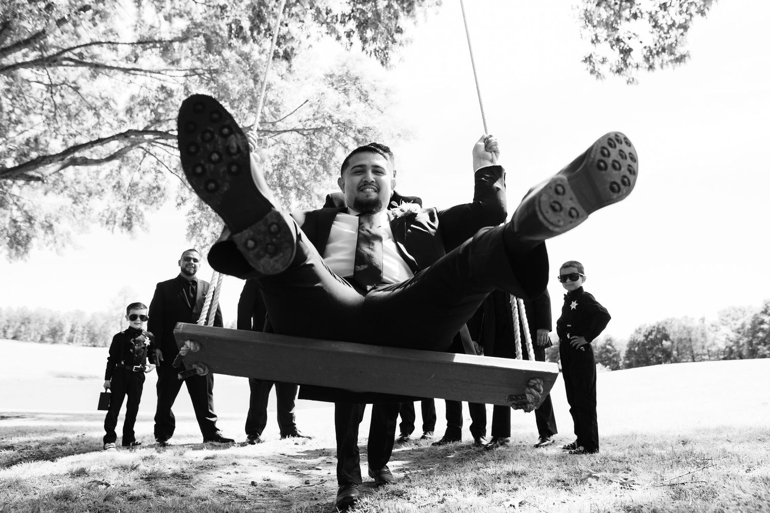 Black and white photo of a joyful groom swinging high on a wooden swing while groomsmen watch in the background.