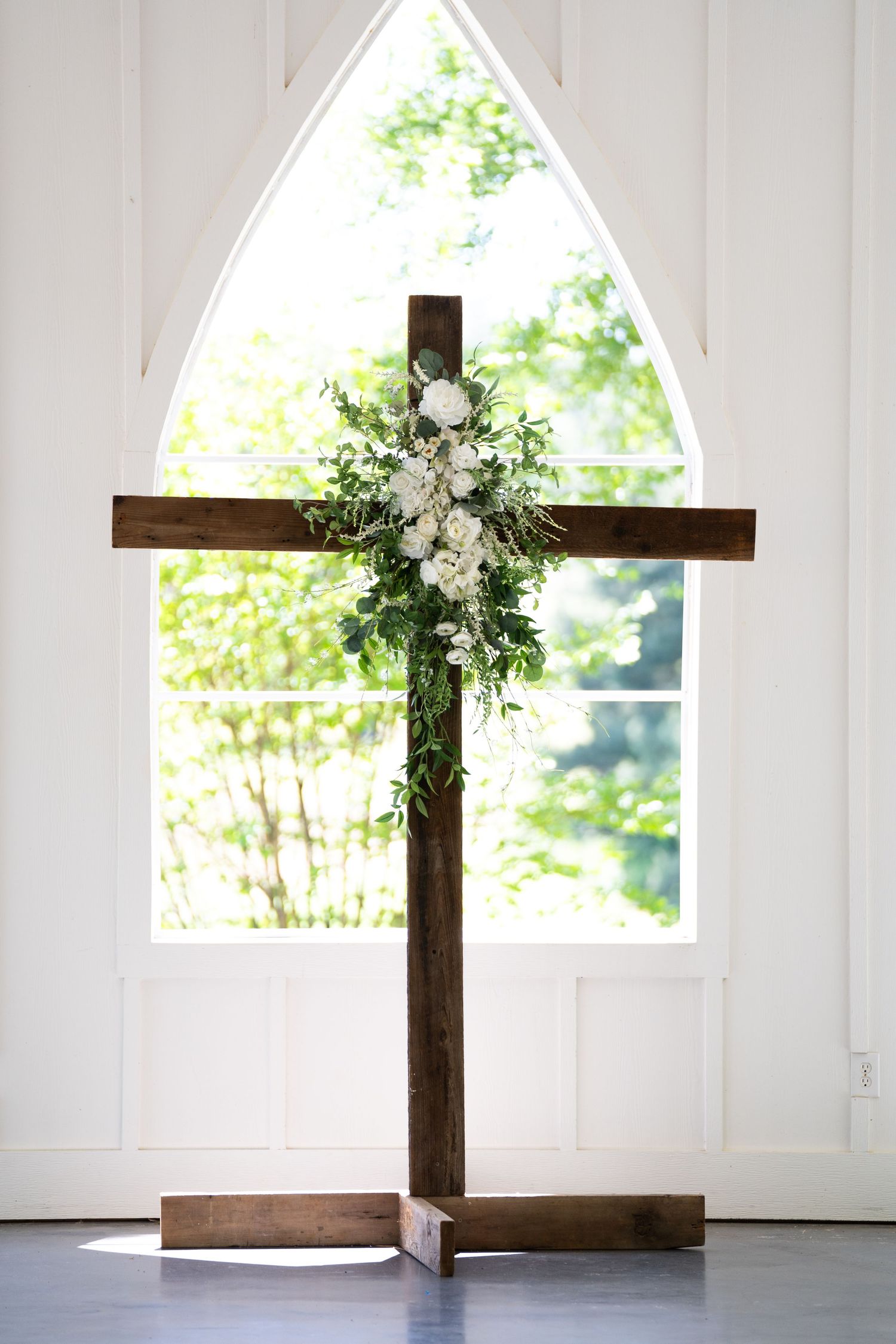 A rustic wooden cross decorated with white flowers and greenery stands in a bright white chapel interior.