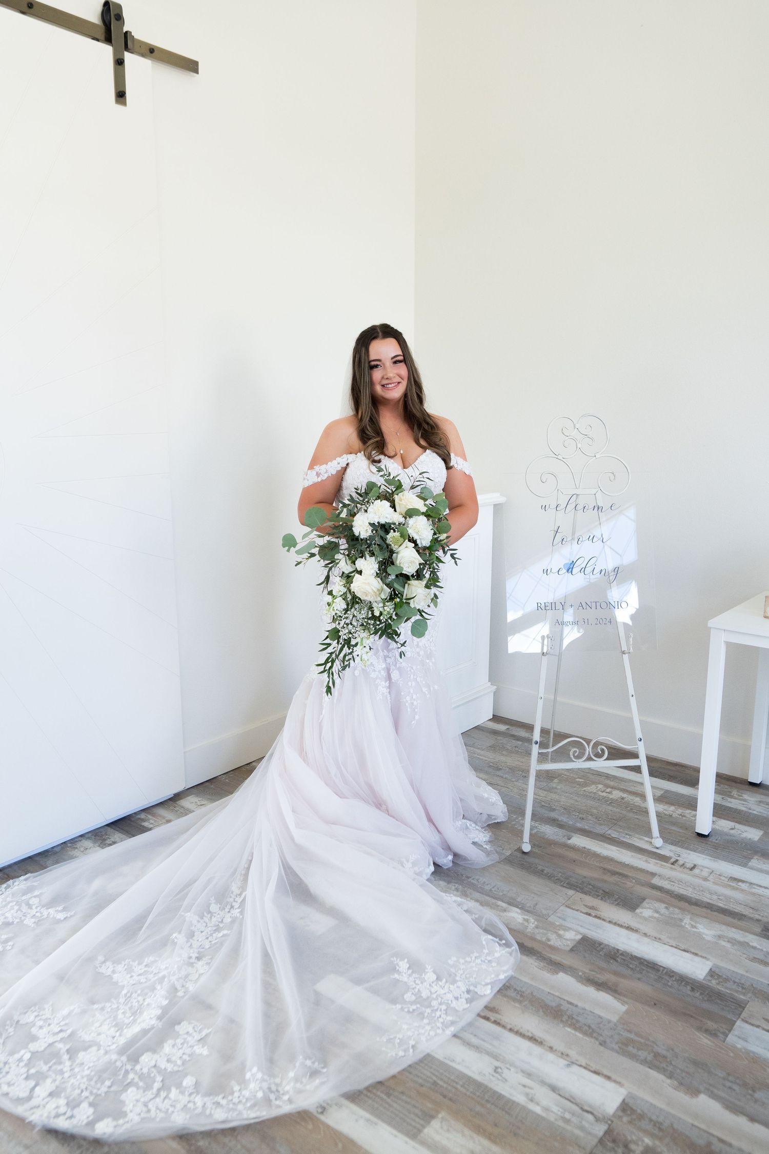 A bride in a white lace wedding gown with a flowing train poses in a bright studio setting with white decor.