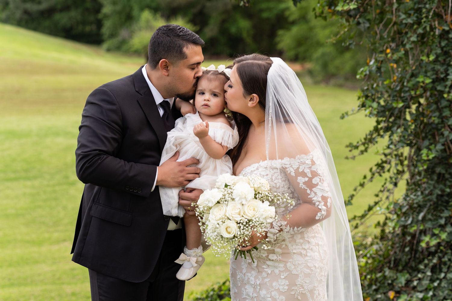 A bride and groom share intimate moments with their baby on a lush green lawn during their wedding celebration.