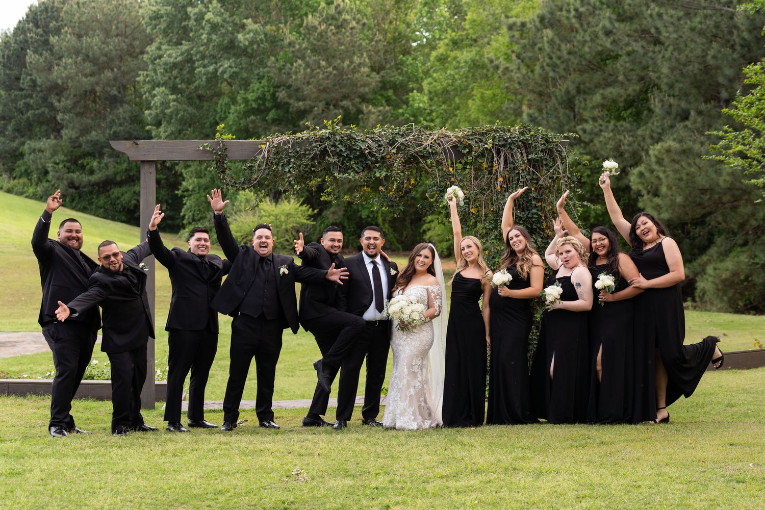Wedding party in black formal attire celebrating joyfully on a green lawn with trees in the background.