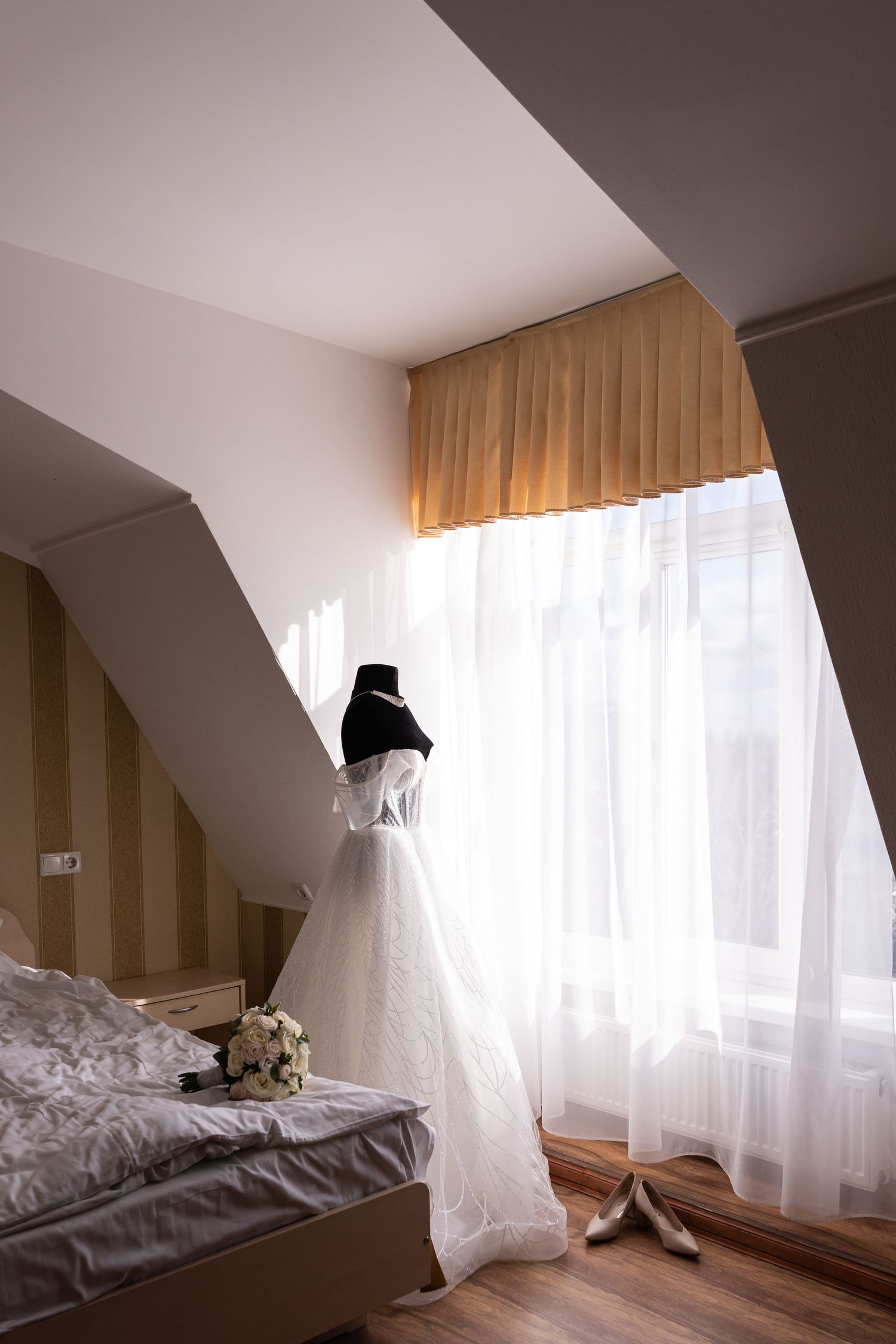 White wedding dress hanging near a window in a modern bedroom with soft natural lighting and wooden accents.