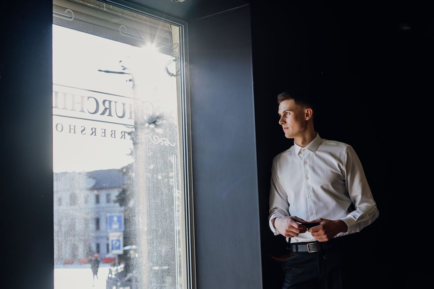 A person in a white shirt stands near a window in a dimly lit room creating a moody silhouette effect.