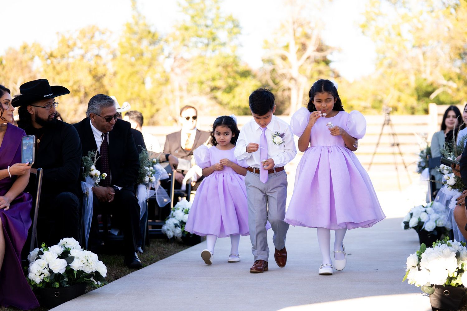 Wedding processional with young attendants in purple dresses walking down outdoor aisle.