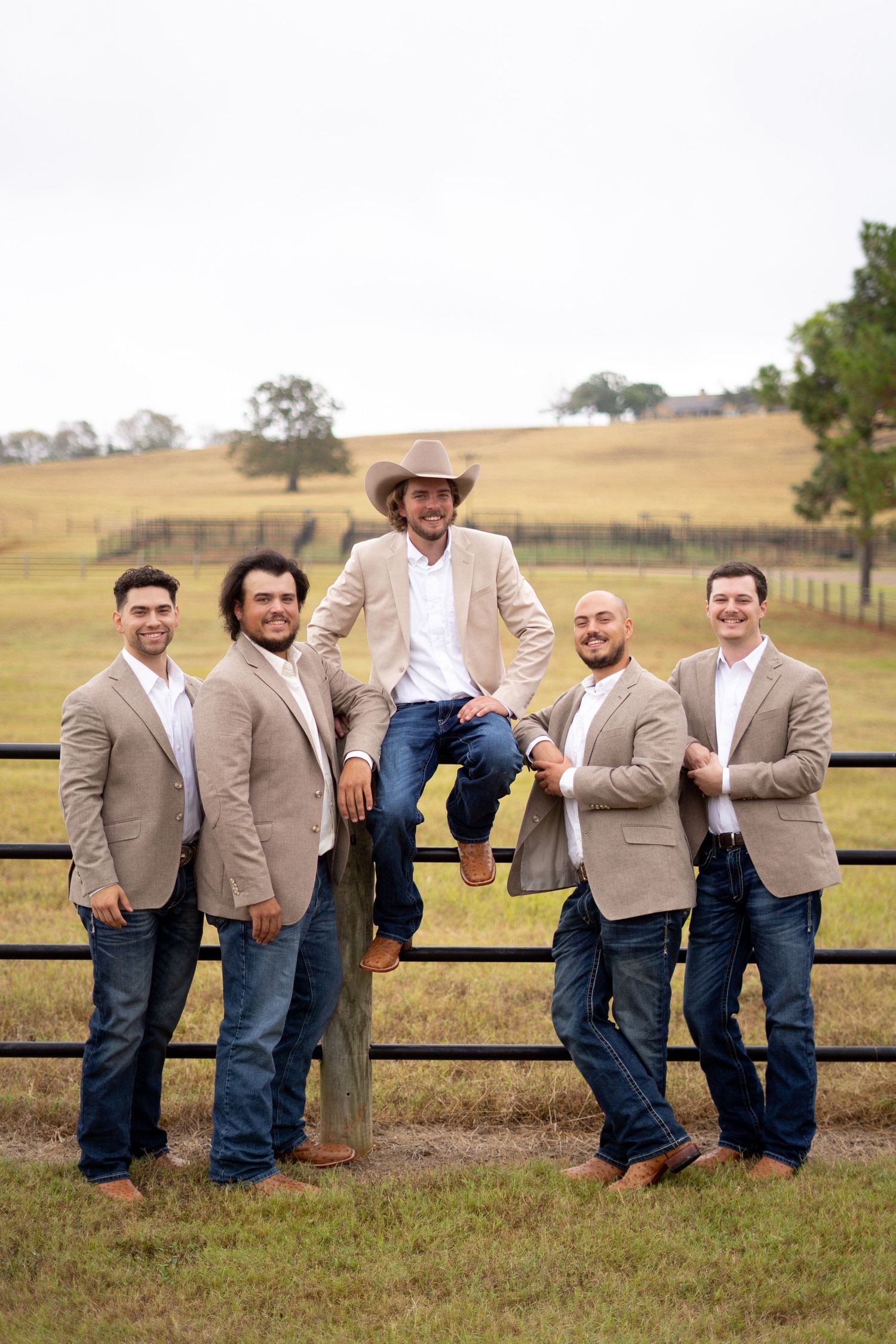 A group of groomsmen in tan suits pose on a wooden fence at a rustic outdoor venue.