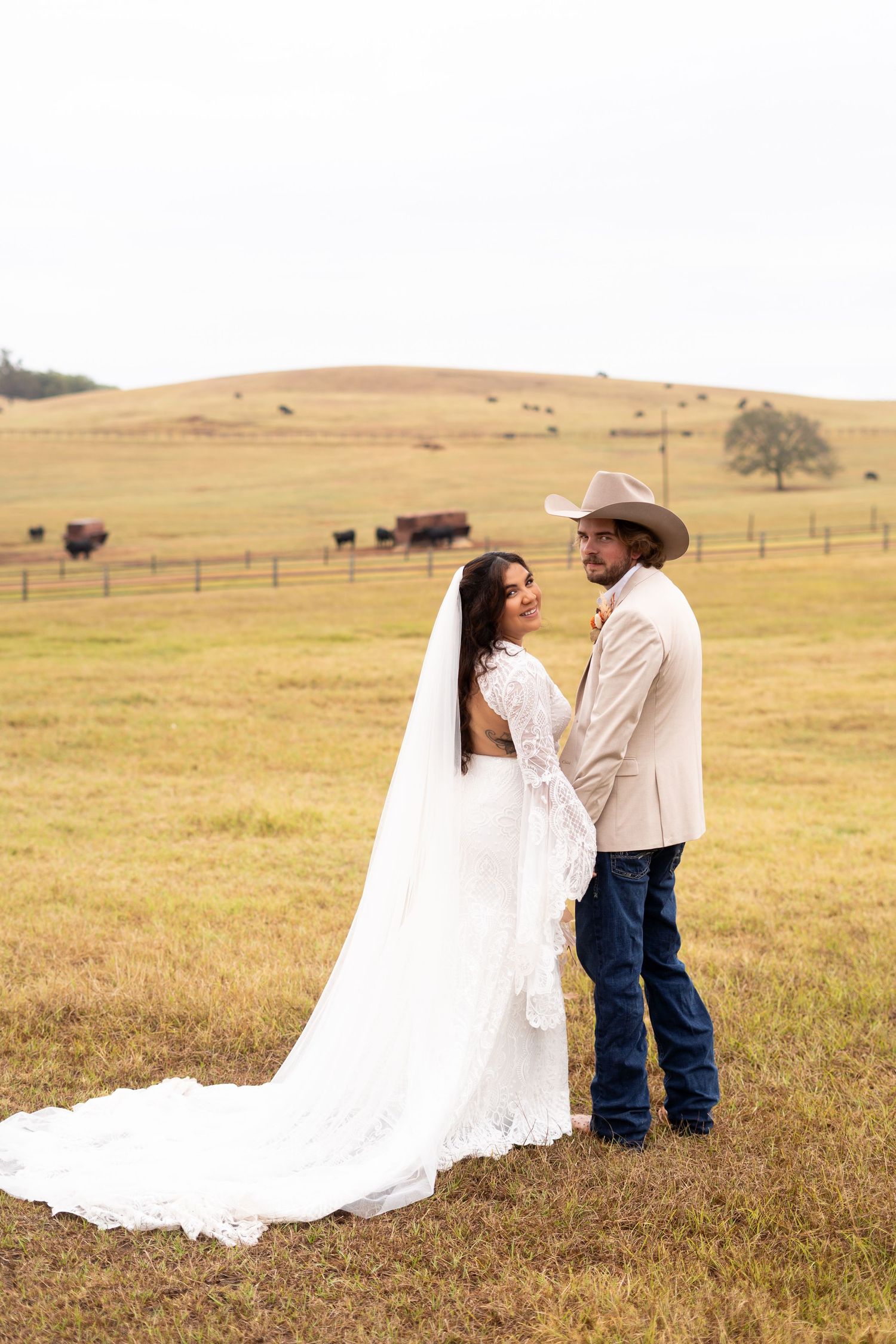 Newlyweds share intimate moment in field with long white wedding dress trailing behind.