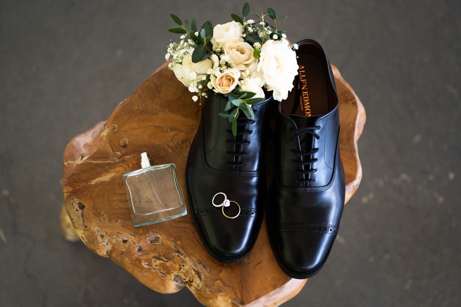 Elegant black leather dress shoes with wedding rings and floral bouquet arranged on brown leather surface.