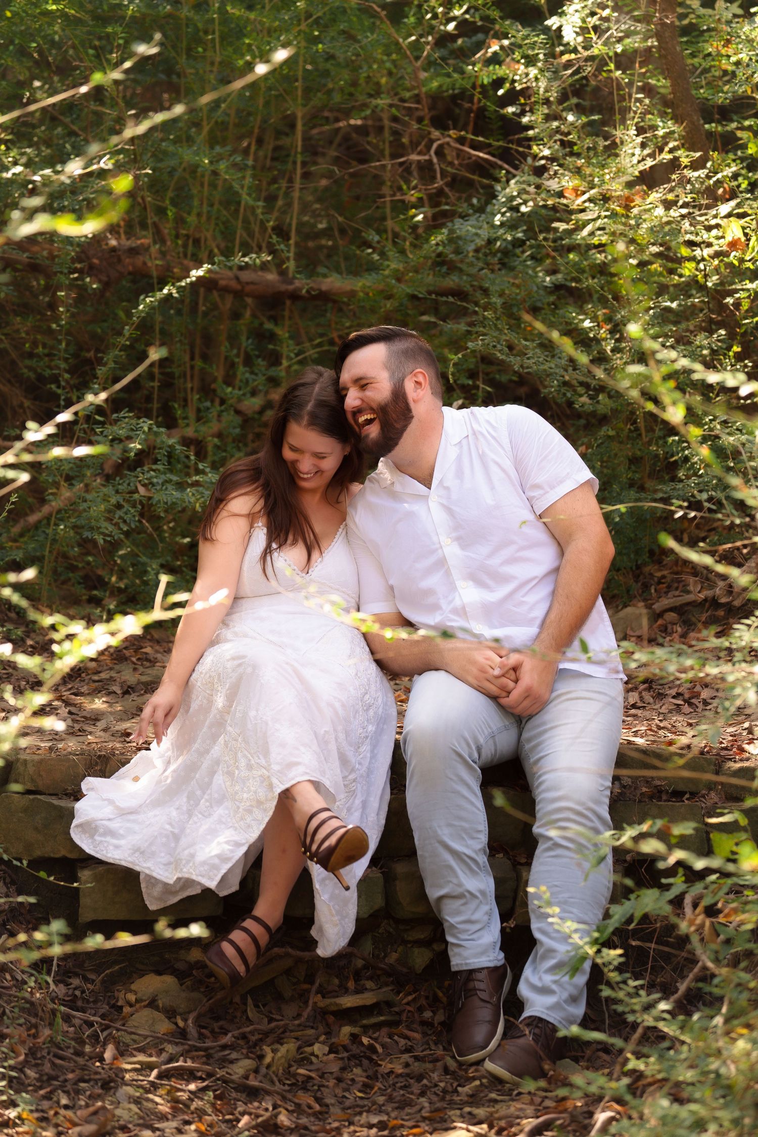 Couple in white clothing shares tender moments while sitting on fallen logs in a wooded setting.